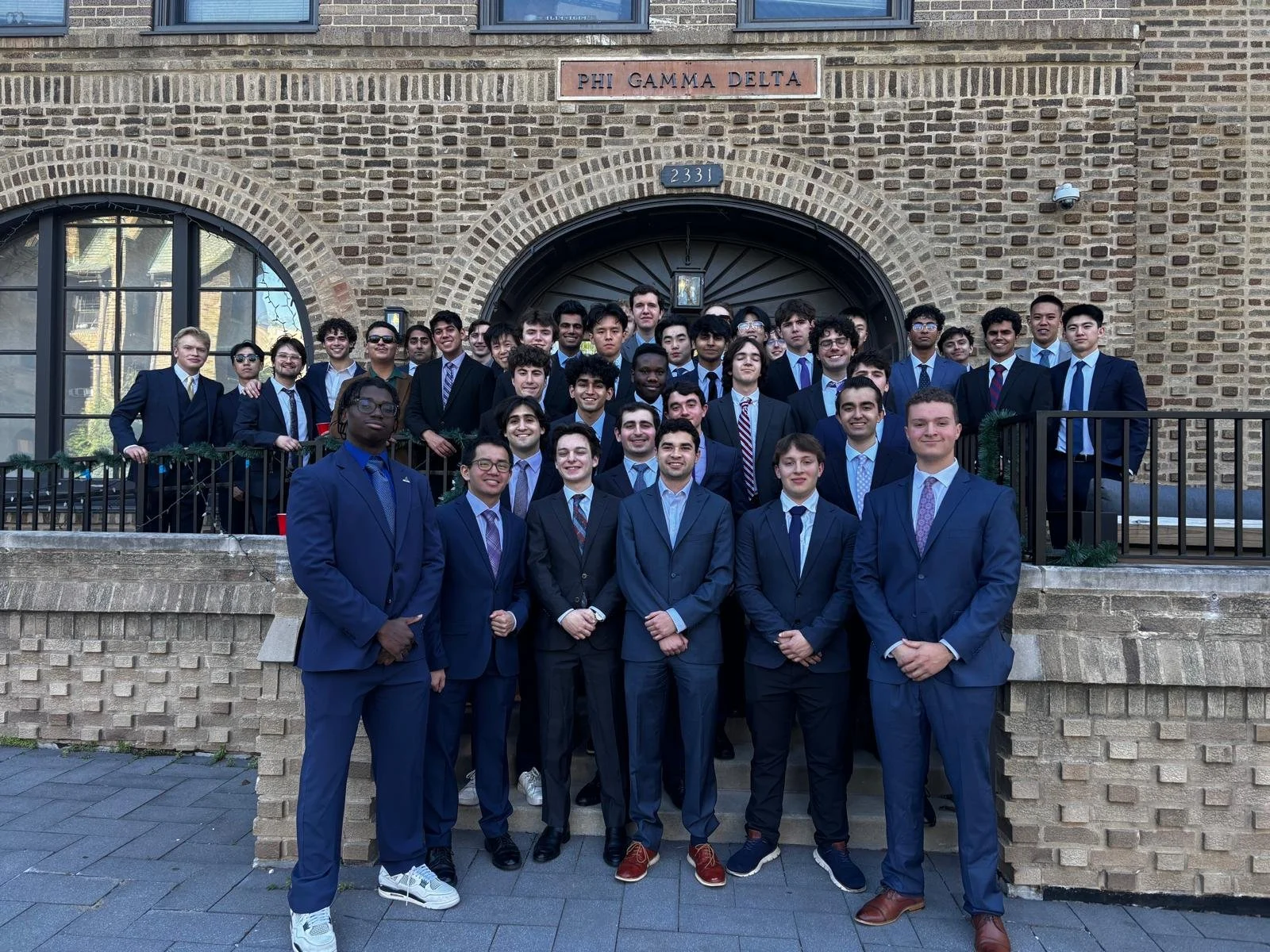 Group of young men in suits standing on steps in front of a building with a sign that says 'Phi Gamma Delta.'