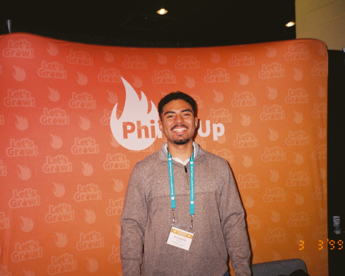 A smiling man stands in front of an orange backdrop with the 'Let's Grow!' logo and a white flame graphic containing the word 'Phil Up' at an event.