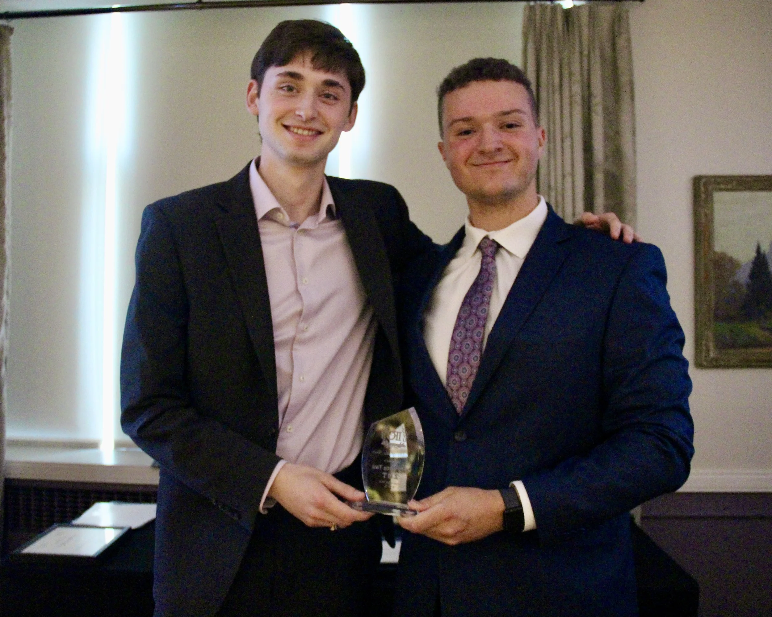 Two young men in formal attire smiling and holding an award together at an indoor event.