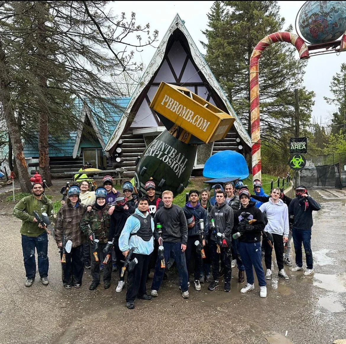 A group of people wearing paintball gear, holding paintball guns, standing in front of a themed amusement park attraction with a large candy cane and a building shaped like a house. There is a sign that reads 'STAGING AREA' with a biohazard symbol.