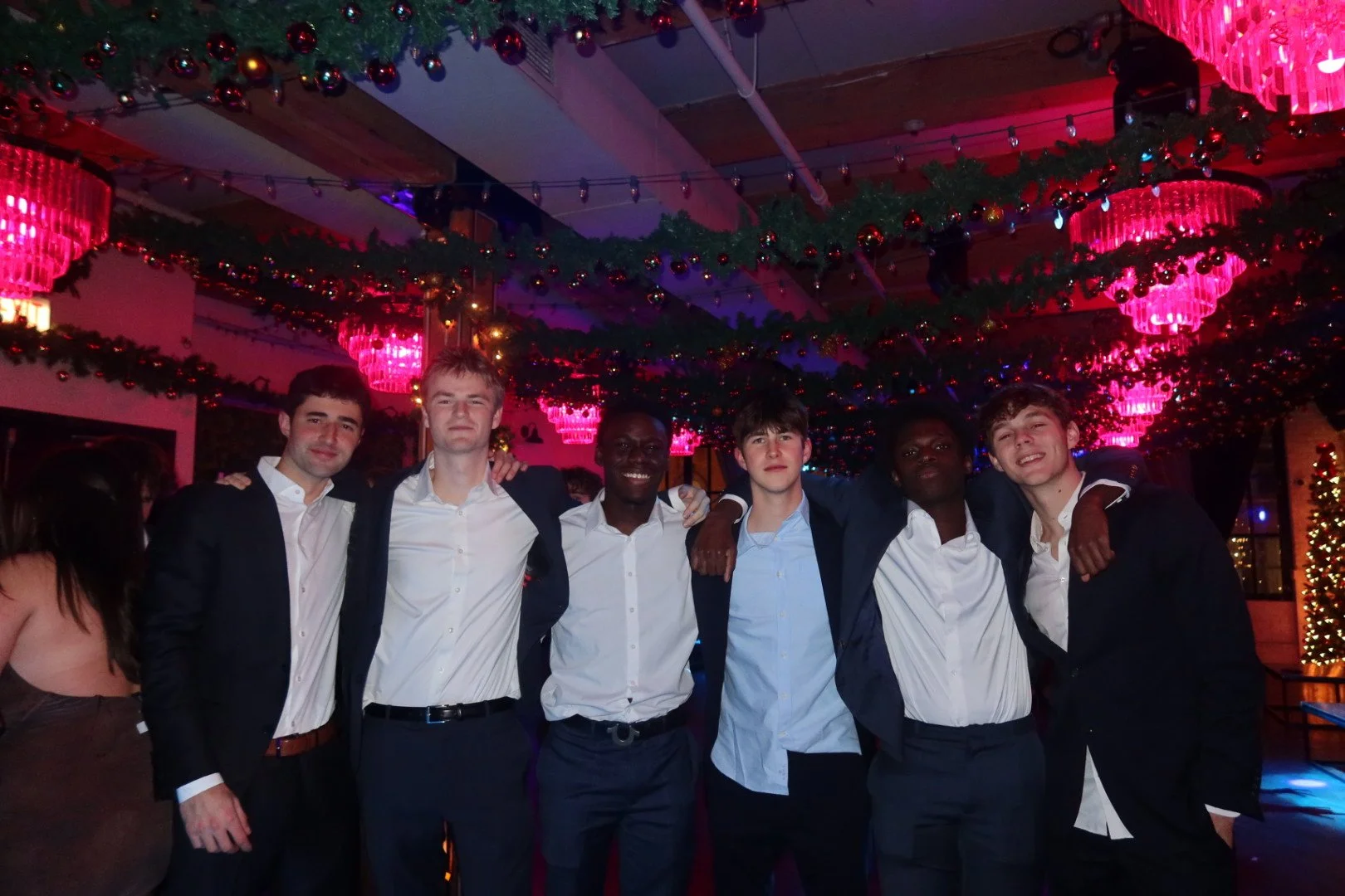 Group of six young men dressed in formal attire standing together at a Christmas party, with festive decorations and pink chandeliers overhead.