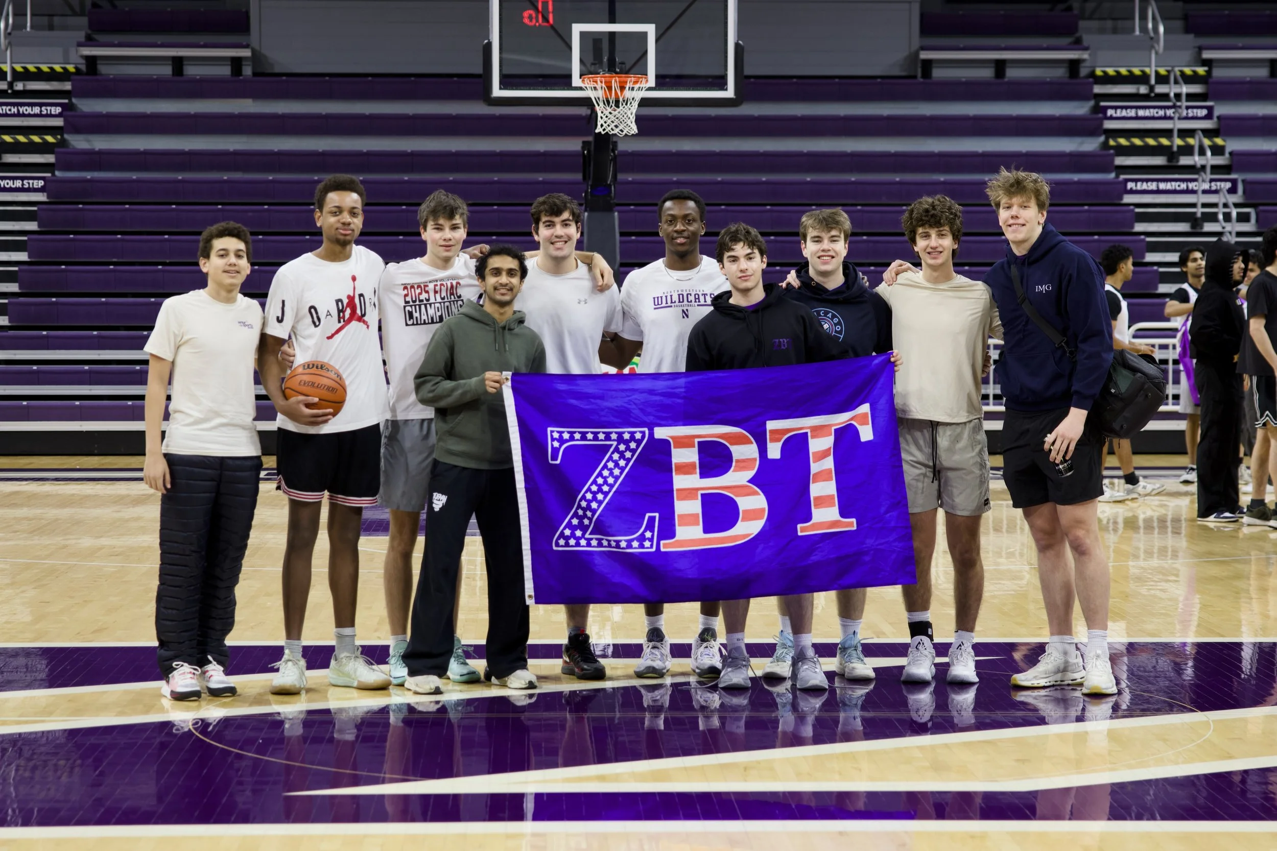 Group of twelve young men standing on a basketball court in a gymnasium, some with their arms around each other, posing for a photo. One person is holding a purple banner with Greek letters, and a couple of individuals are holding a basketball. The gym has purple bleachers and signs that say 'Please Watch Your Step'.