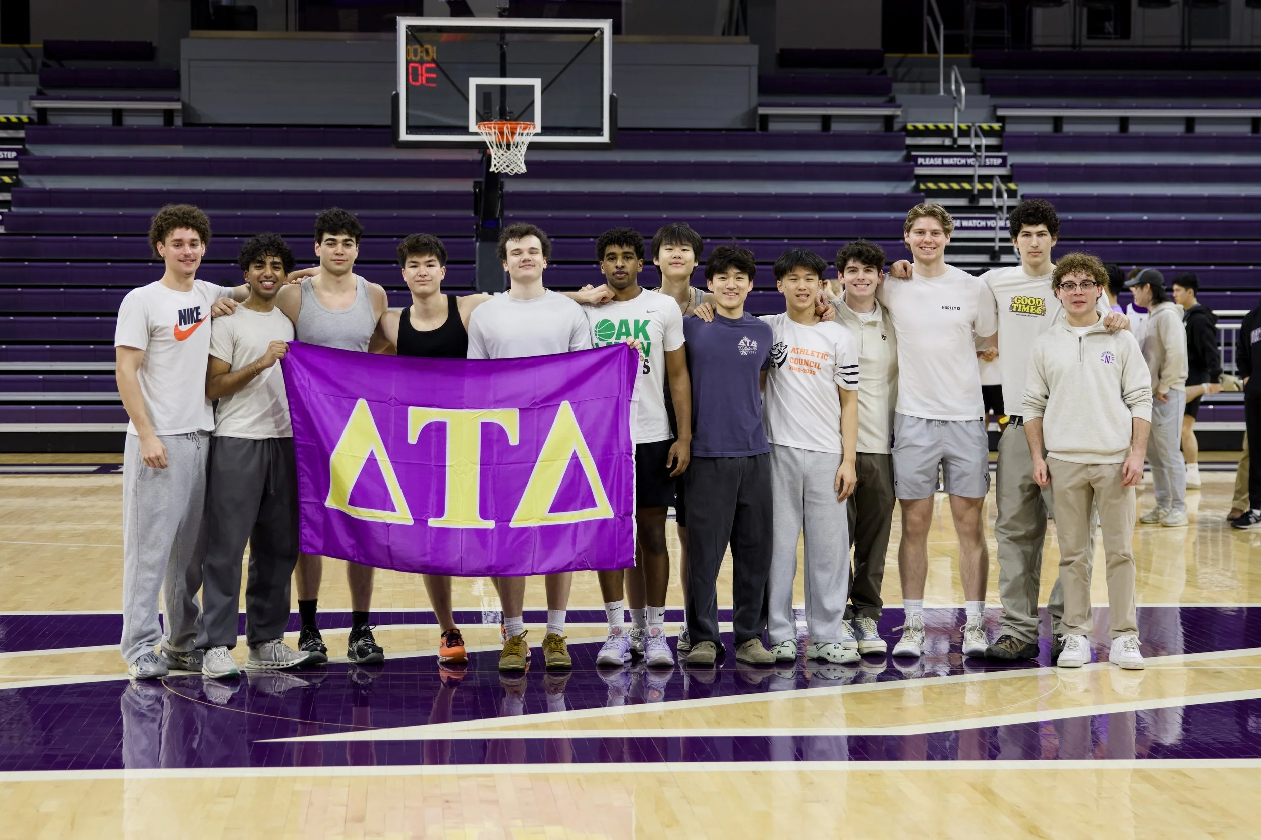 Group of young male athletes standing on a basketball court inside an arena, holding a purple banner with Greek letters, smiling, with a basketball hoop in the background.