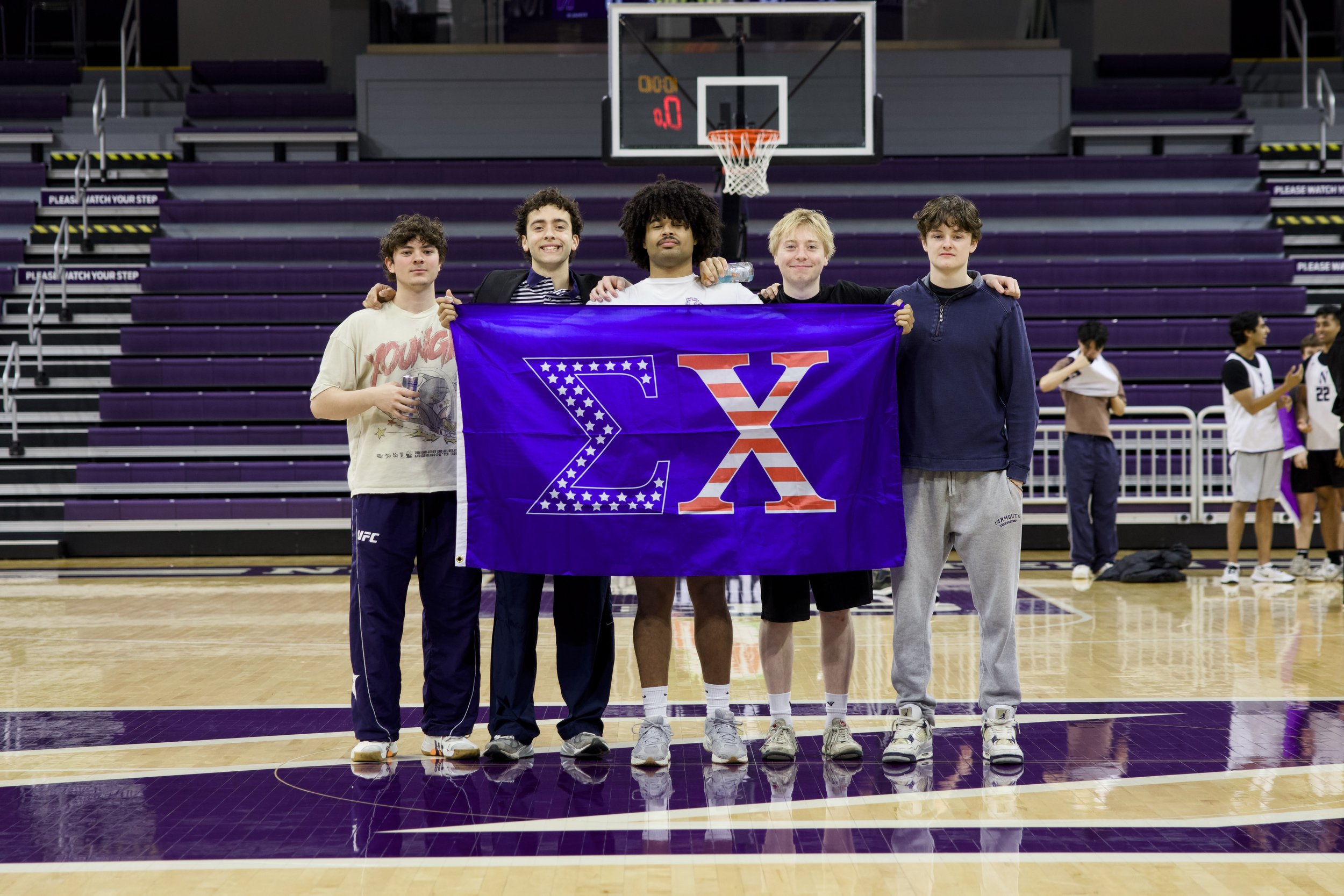 Five young men standing side by side on a basketball court indoors, holding a large purple banner with a Greek letter Delta and American flag design, smiling at the camera, with a basketball hoop behind them.