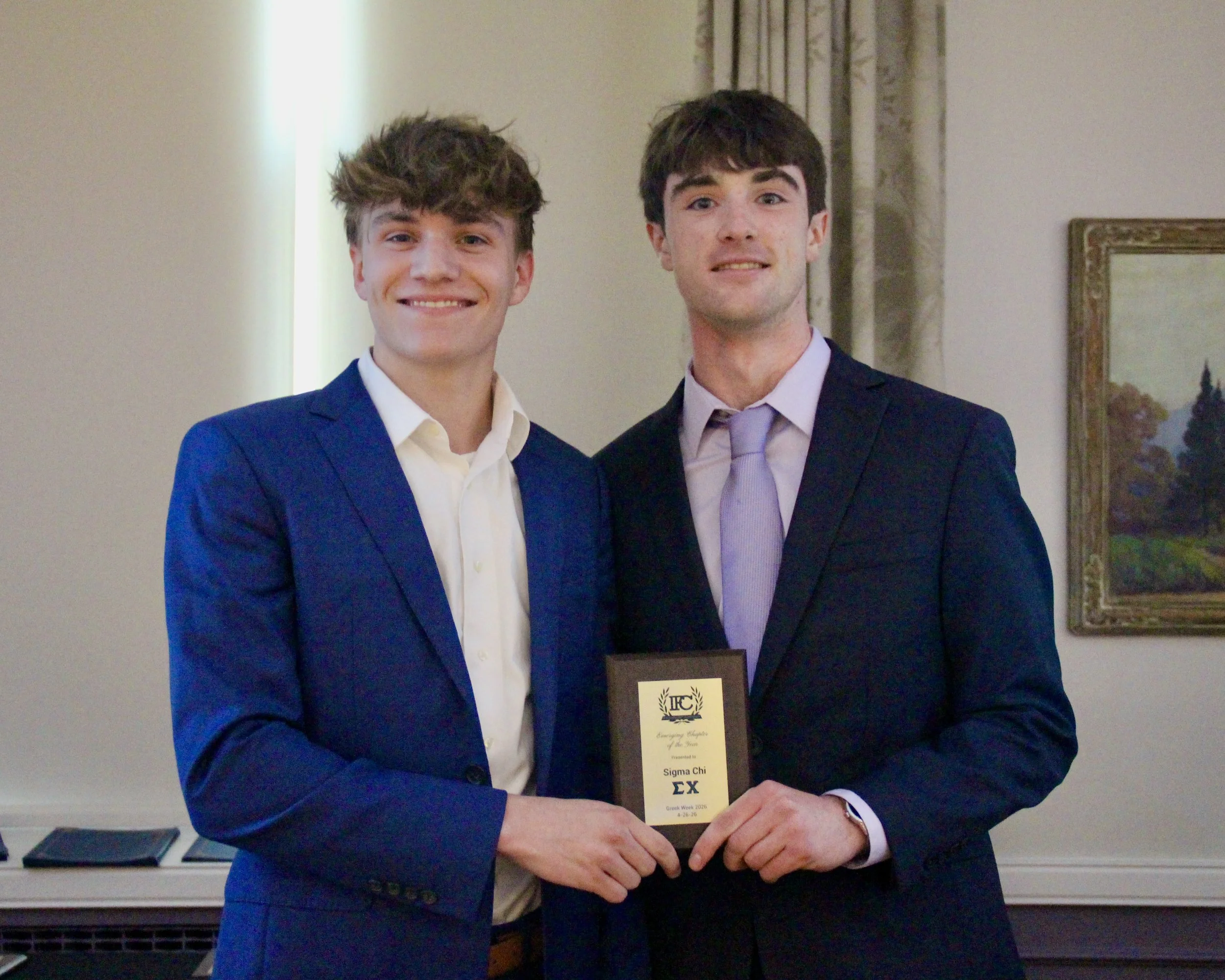 Two young men in suits are smiling and shaking hands at an indoor event. One is holding a Sigma Chi fraternity award.