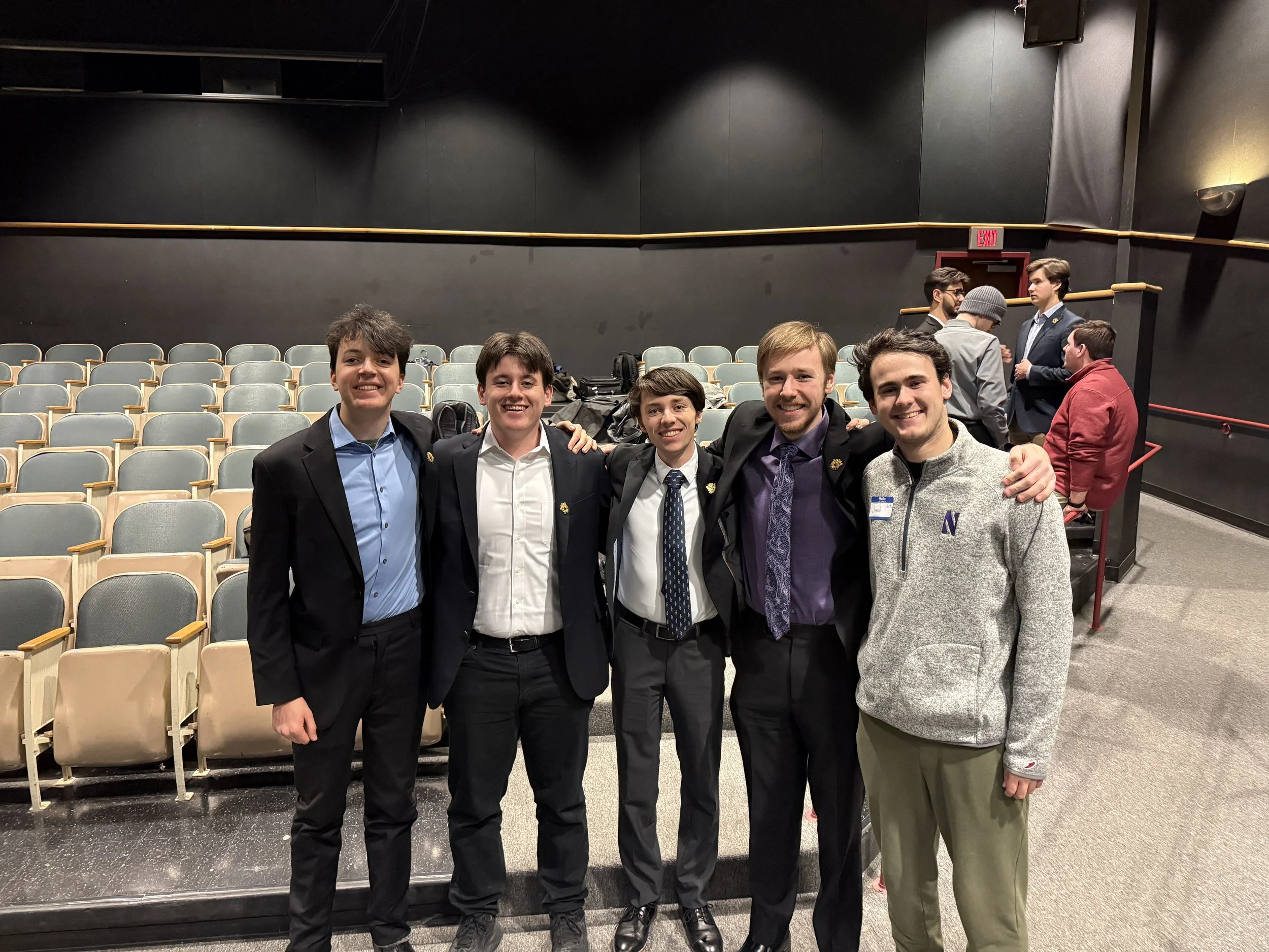 Group of five young men standing together in theater seats, smiling, with four people in the background engaged in conversations.