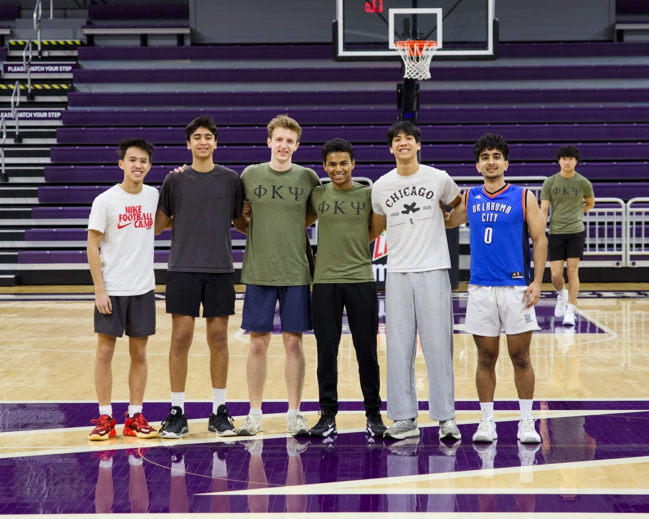 Group of seven young men standing on a basketball court in an indoor stadium. They are smiling and posing for the photo with purple seating in the background. Some are wearing sports t-shirts, and one is in a basketball jersey. The court has purple and yellow markings, and there is a basketball hoop above them.