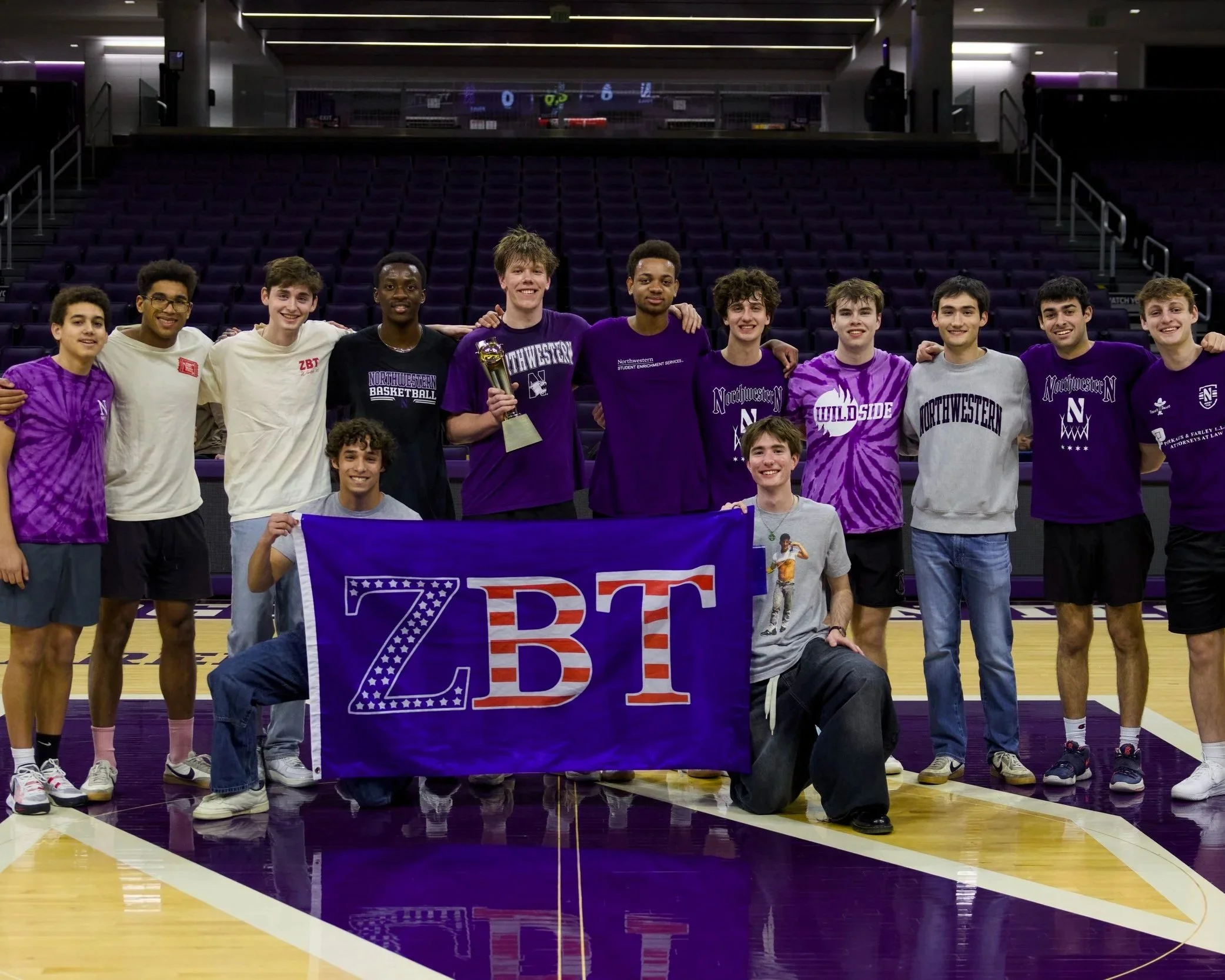 A group of young male basketball players standing together on a basketball court, holding a banner and a trophy, smiling for a photo.