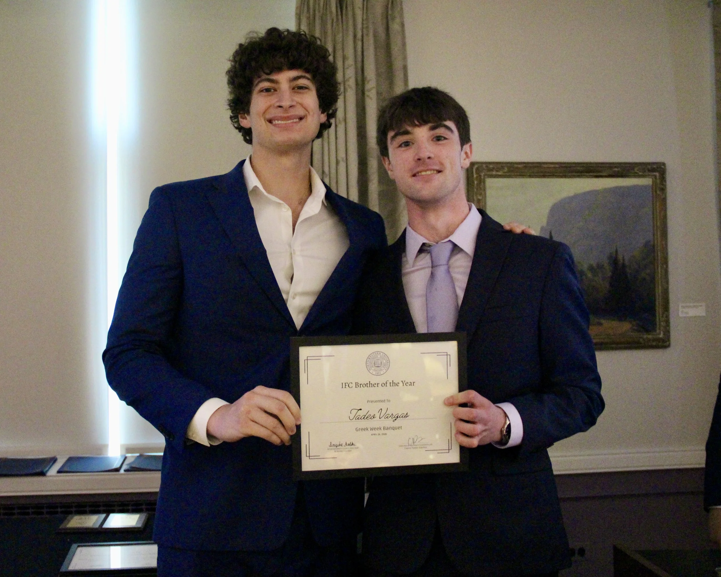Two men in business suits standing together, smiling, one holding a certificate that reads "IFC Brother of the Year." They are indoors, with a painting and some books or awards on a table behind them.