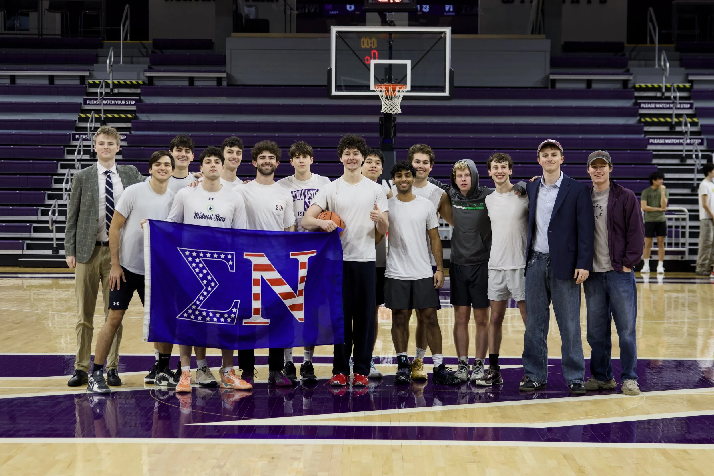 A group of young men and women in basketball uniforms and casual clothing are standing on a basketball court inside a gym, holding a large blue flag with a Greek letter symbol and stars. They are smiling and posing for the photo, with purple bleachers and a basketball hoop visible in the background.