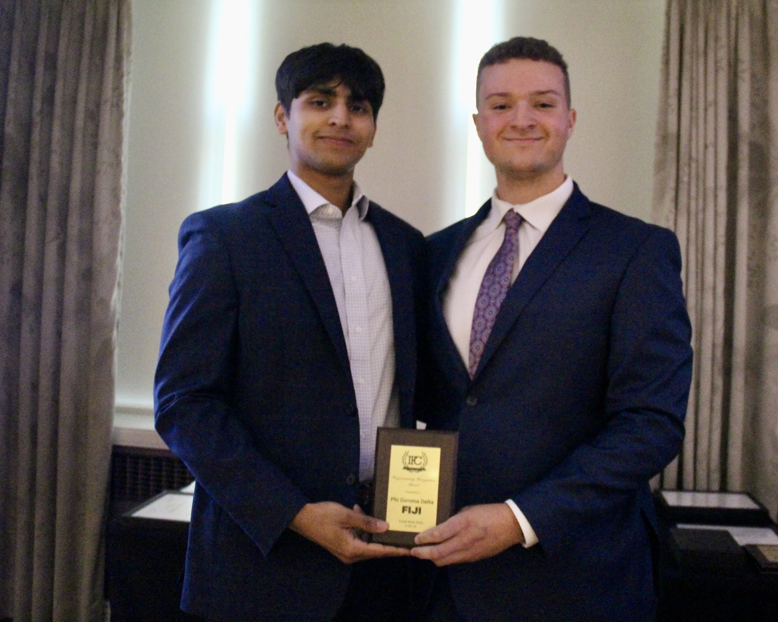 Two men in formal suits standing close together, smiling for a photo indoors. The man on the right is holding a yellow certificate or award.