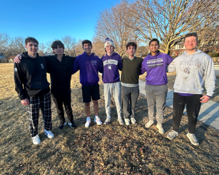 Seven young men standing outdoors on a sunny day, with a large tree and a sidewalk in the background. They are dressed casually, some wearing hoodies and sweatshirts, and are smiling with arms around each other.