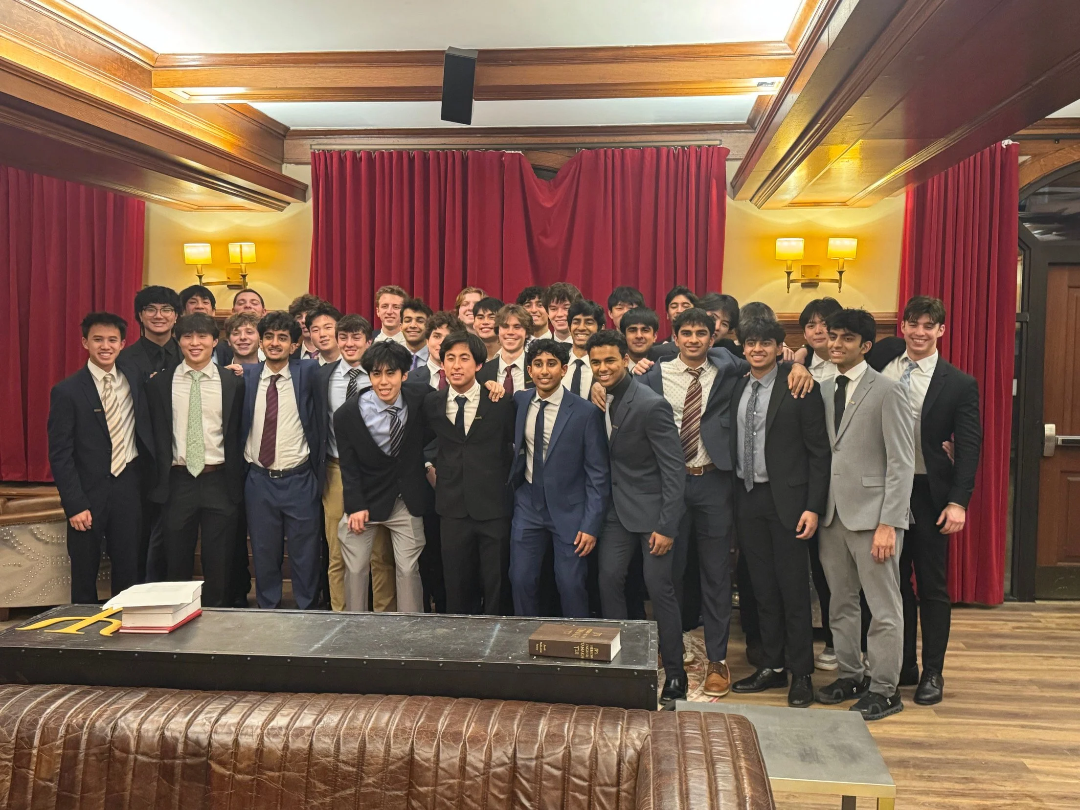 Group of young men in suits standing in front of red curtains with warm lighting, smiling for a photo in an elegant room.
