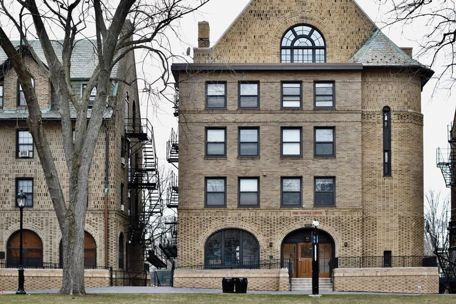 A historic brick apartment building with fire escapes on either side and a small front yard with a tree and lamp post.