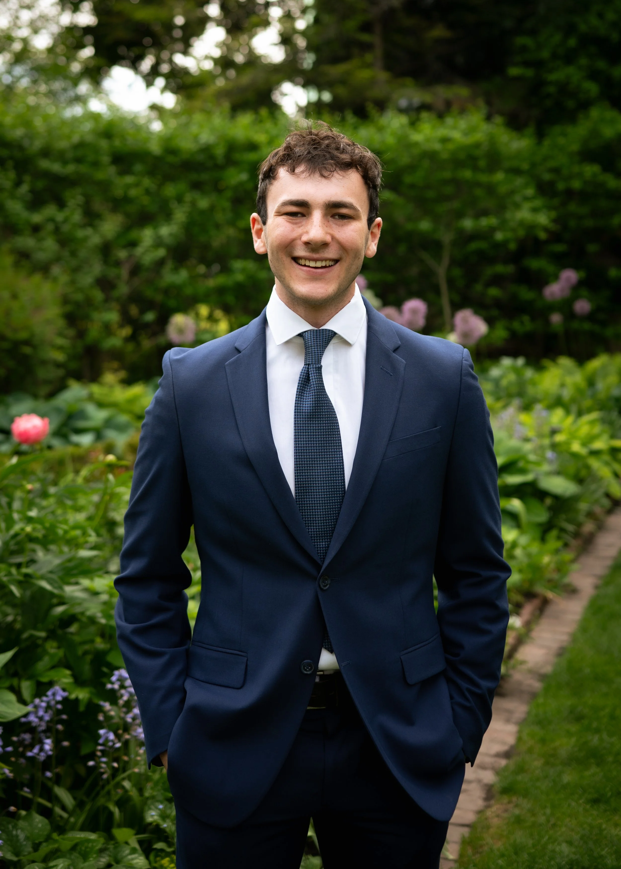 A young man in a navy suit with a white shirt and blue tie smiling outdoors in a garden with green foliage and pink flowers.