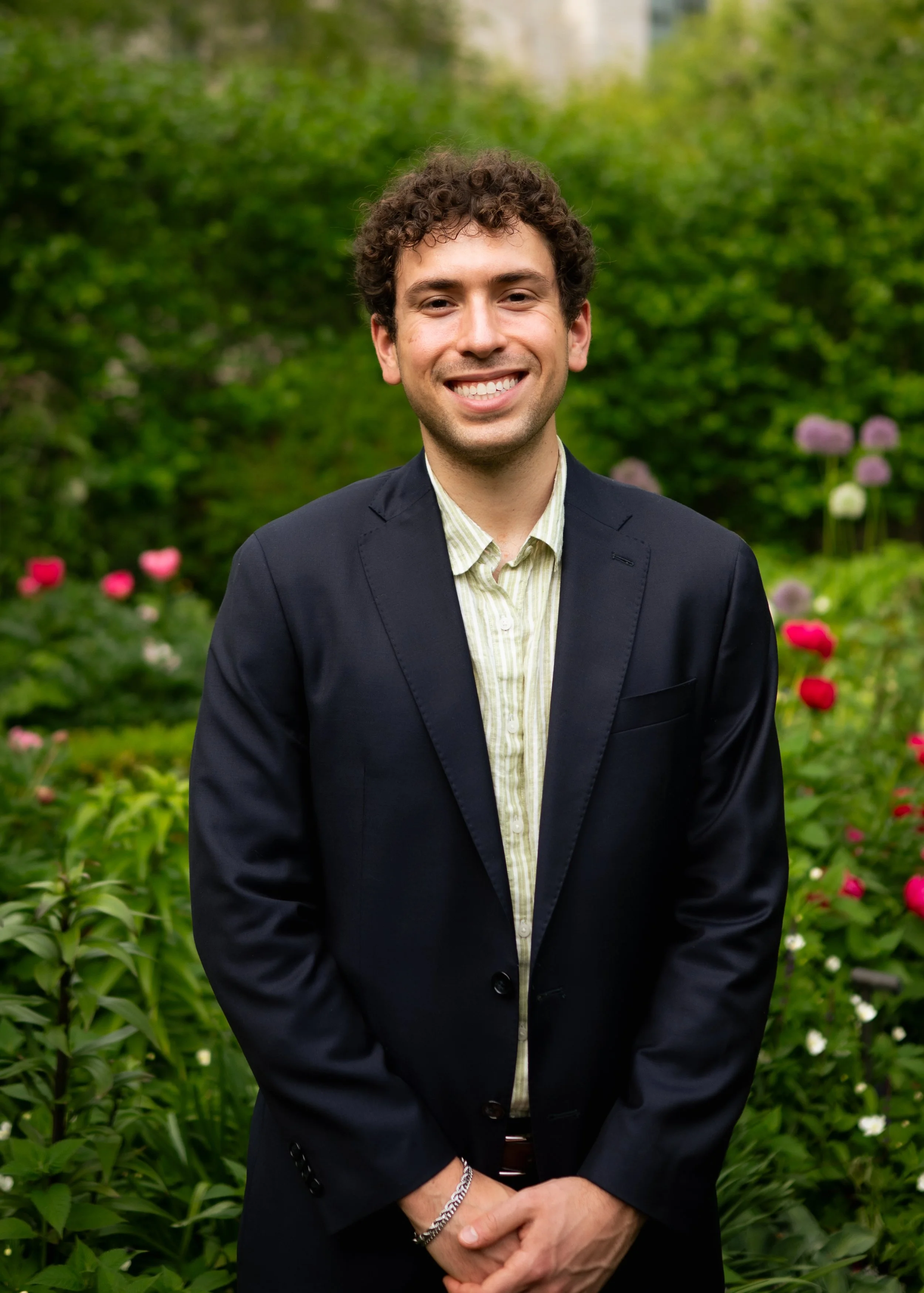 A young man with curly brown hair, wearing a dark suit jacket and a light-colored shirt, smiling and standing outdoors in front of green bushes and blooming flowers.