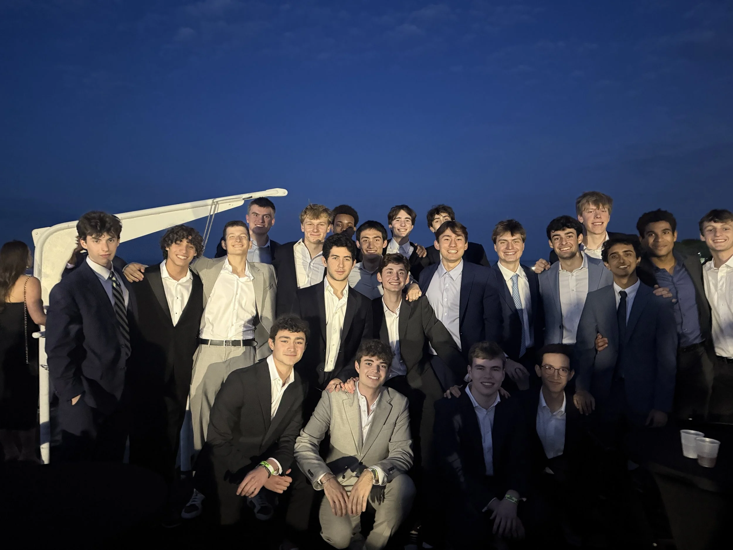 Group of young men dressed in formal suits smiling and posing for a photo at dusk, outdoors with a darkening sky in the background.