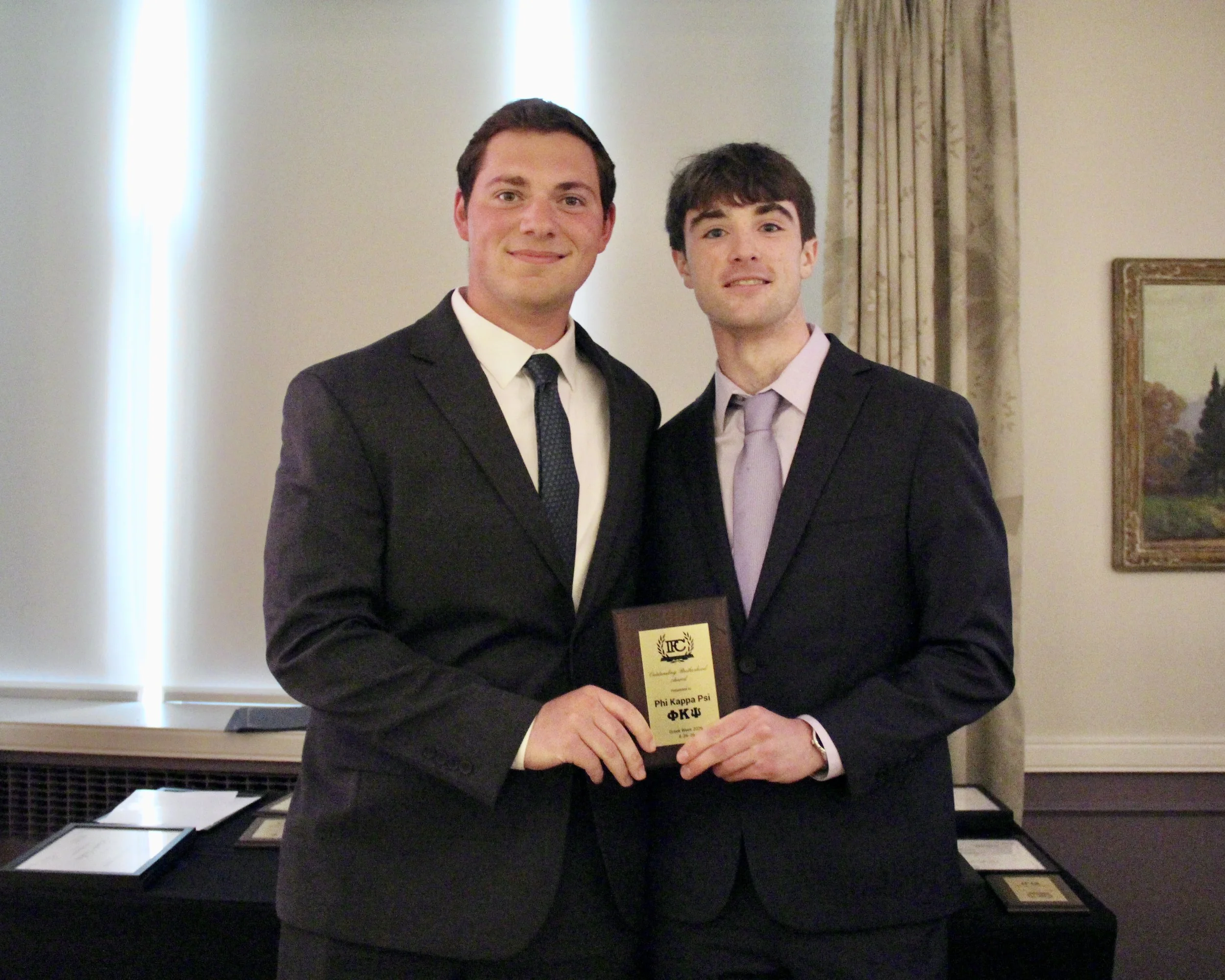 Two young men in formal suits standing together at an indoor event, one holding a Phi Kappa Psi fraternity certificate or award.