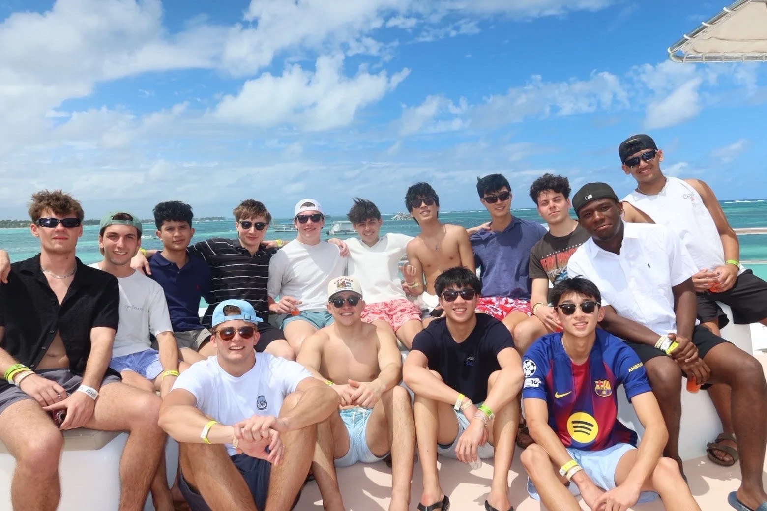 Group of young men enjoying a boat trip on the ocean, some wearing swim trunks and sunglasses with clear blue skies and boat in the background.