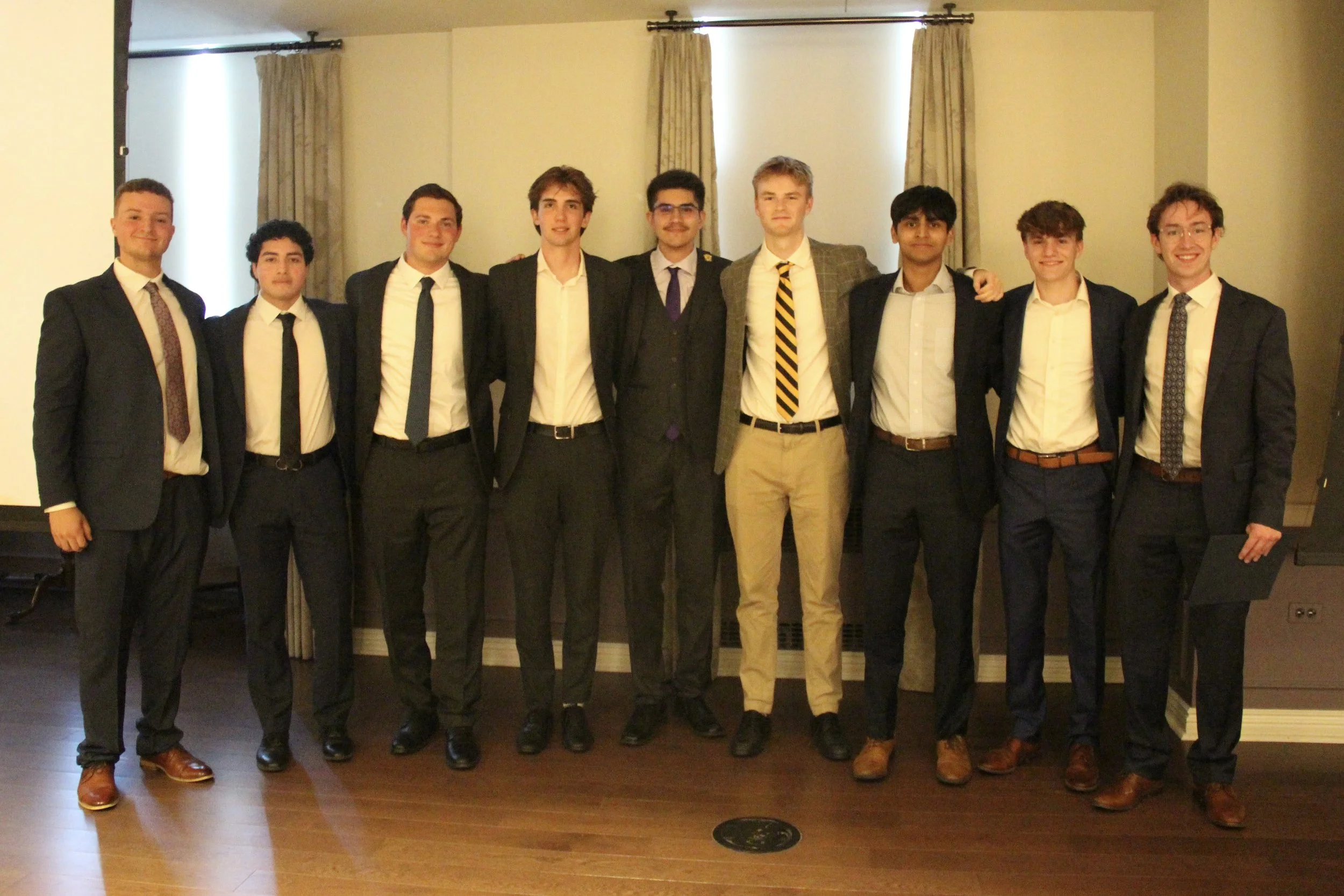 Group of nine young men dressed in formal suits standing in a row indoors.