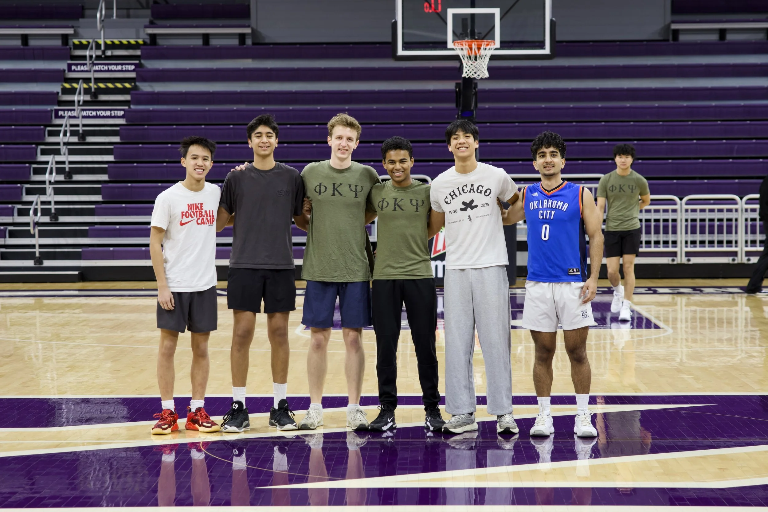 Group of six young men standing in a basketball court, smiling at the camera. They are wearing casual sportswear, with some in t-shirts and others in basketball jerseys. The court has purple and wood-colored flooring, and there are purple bleachers and basketball hoops in the background.
