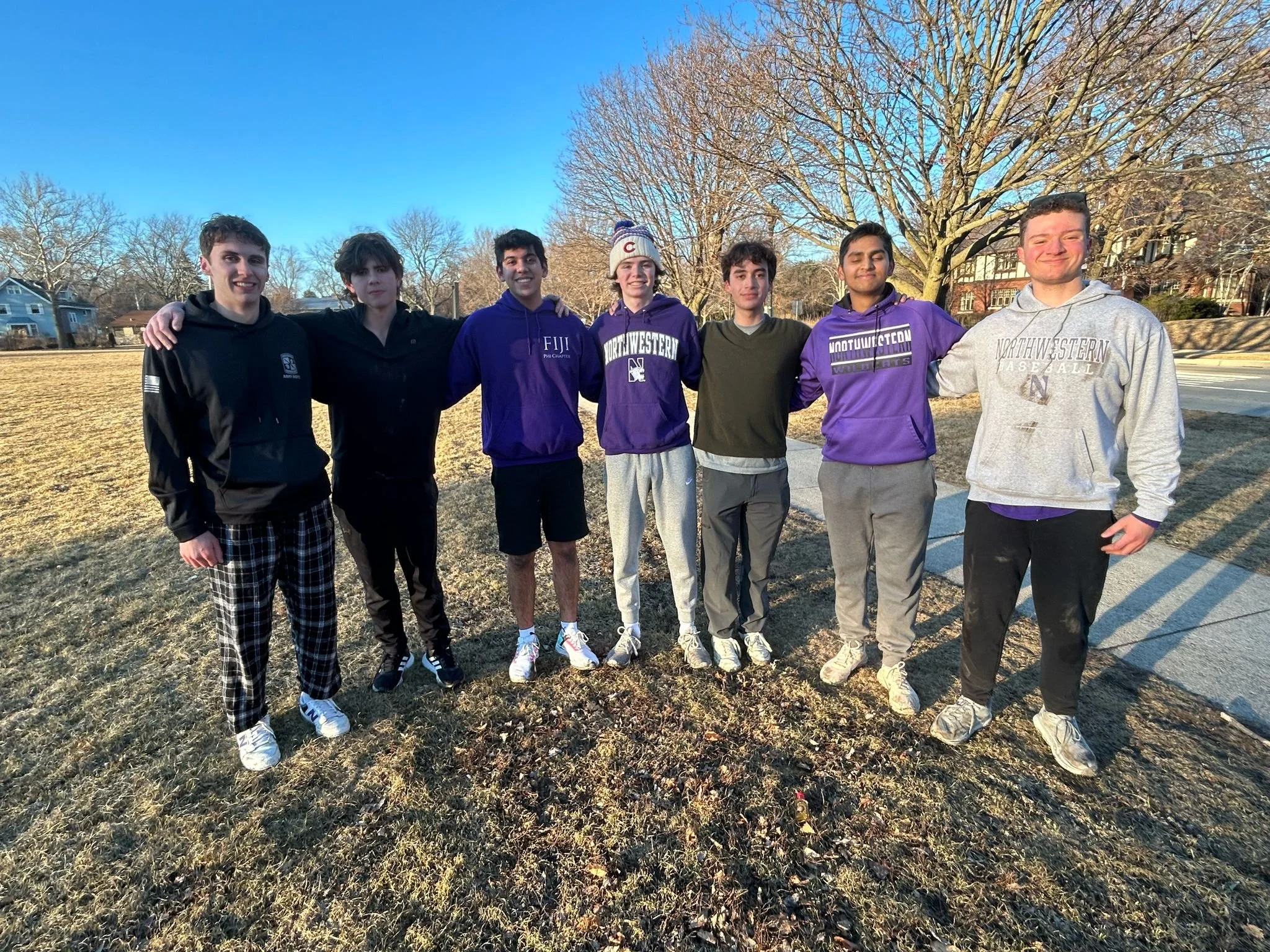 A group of seven young men standing outdoors on a grassy area during winter or early spring, with trees without leaves and houses in the background, smiling and posing for the photo.