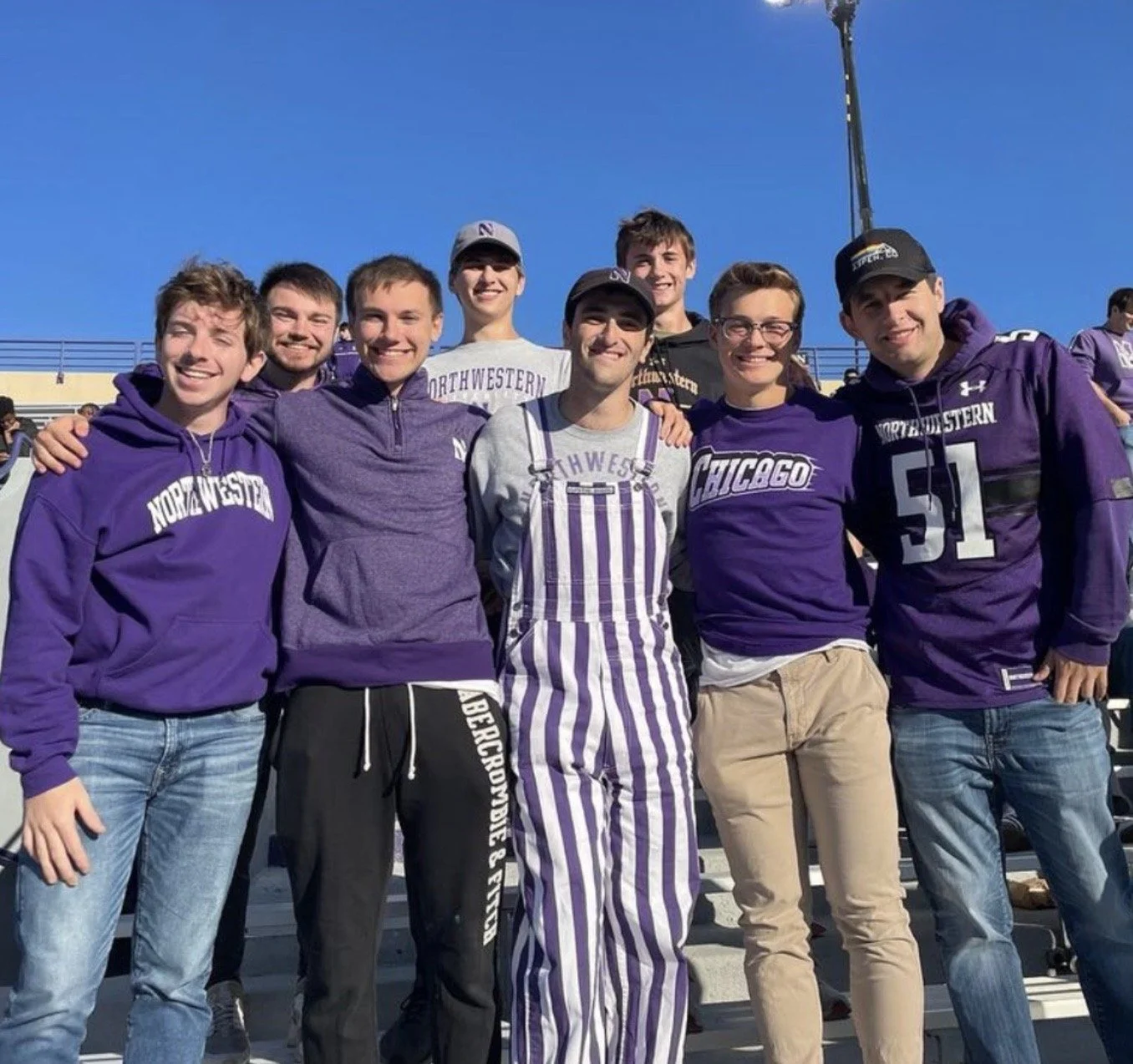 A group of nine young adults standing on bleachers at a sports stadium, wearing Northwestern University and Chicago sports apparel, smiling at the camera on a sunny day.