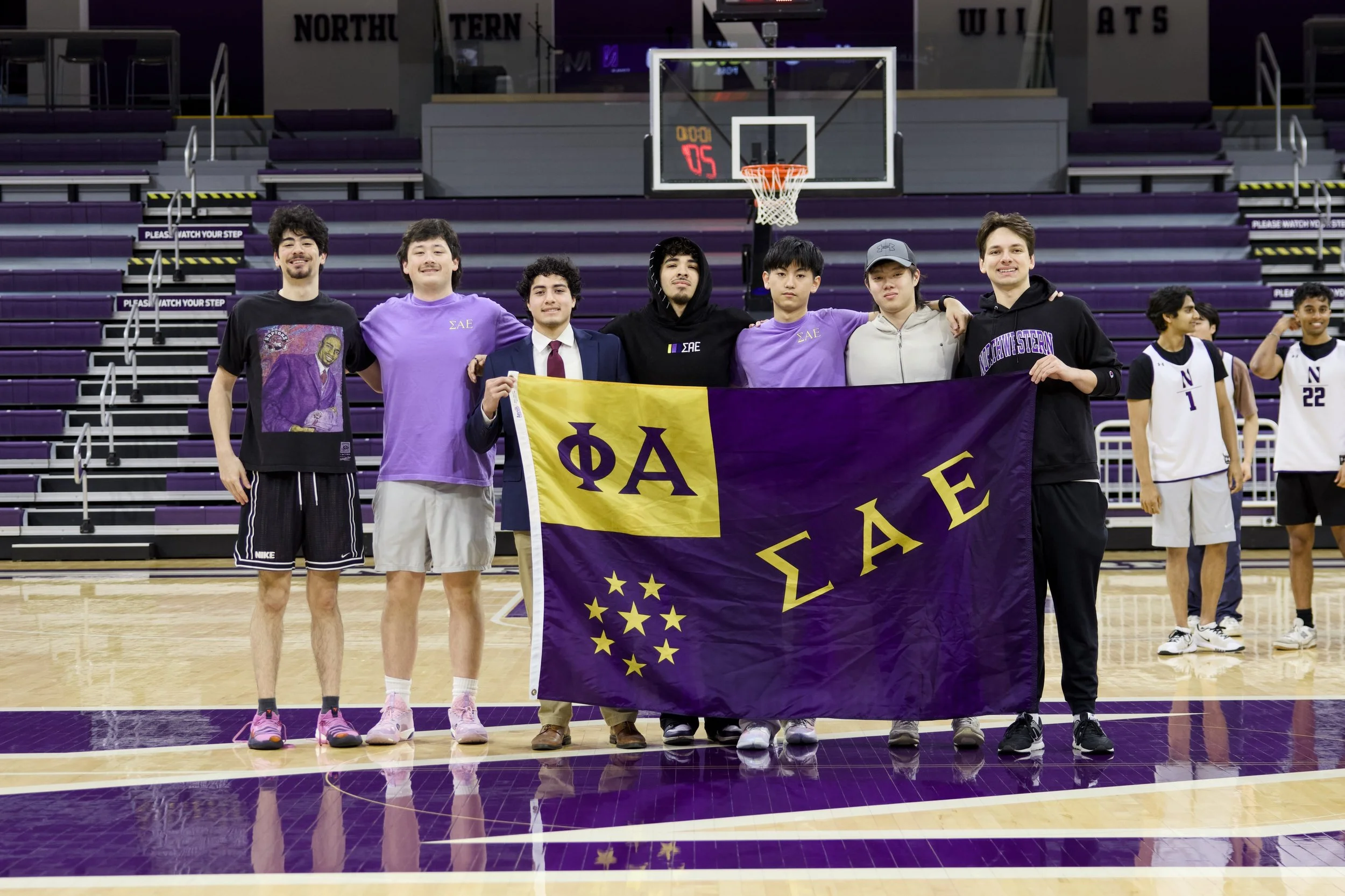 A group of young men standing on a basketball court, holding a purple and yellow sorority banner with Greek letters. They are smiling and posing for the photo, with a few wearing purple shirts and others in different athletic attire. The gymnasium has purple seating and a basketball hoop in the background.