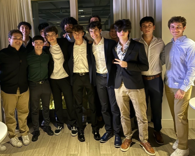 Group of young men dressed in formal and semi-formal attire standing together indoors for a photo, some making peace signs, with a window and curtains in the background.
