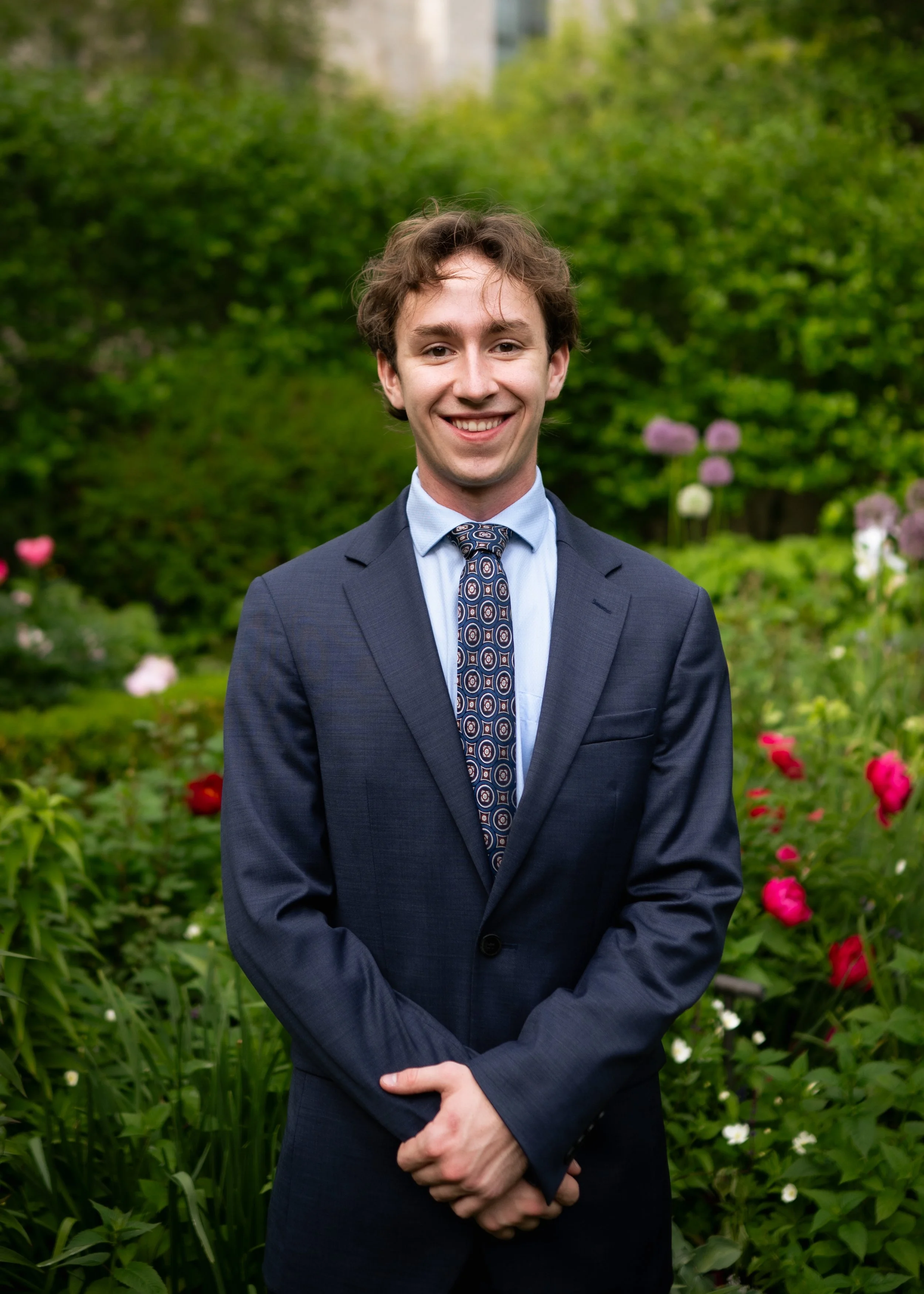 A young man in a dark suit with a patterned tie, smiling and standing outdoors in a garden with colorful flowers and green bushes.