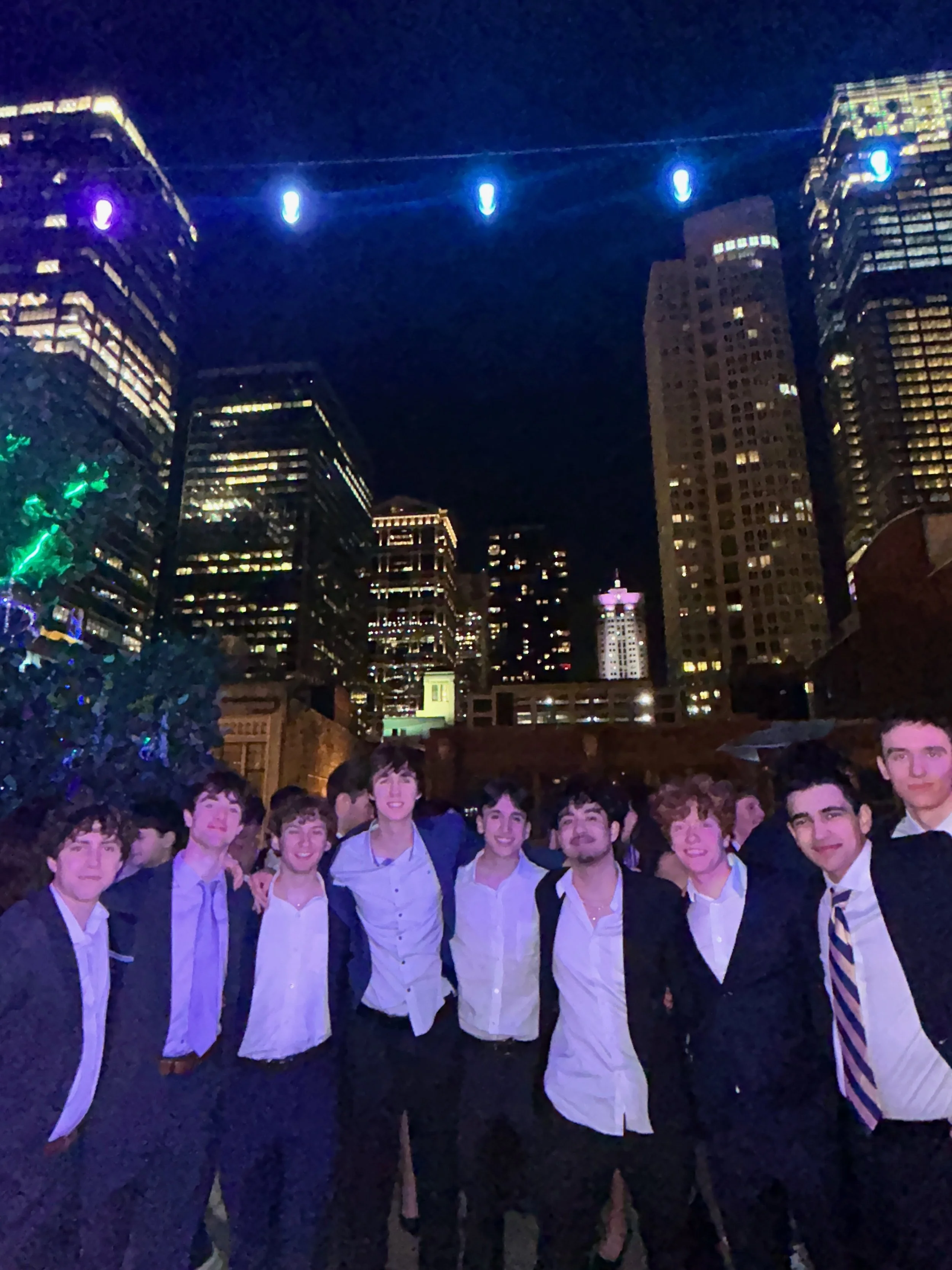 A group of young men dressed in white shirts, suits, and ties standing together outdoors at night in front of a city skyline with tall lit-up buildings and colorful decorative lights.