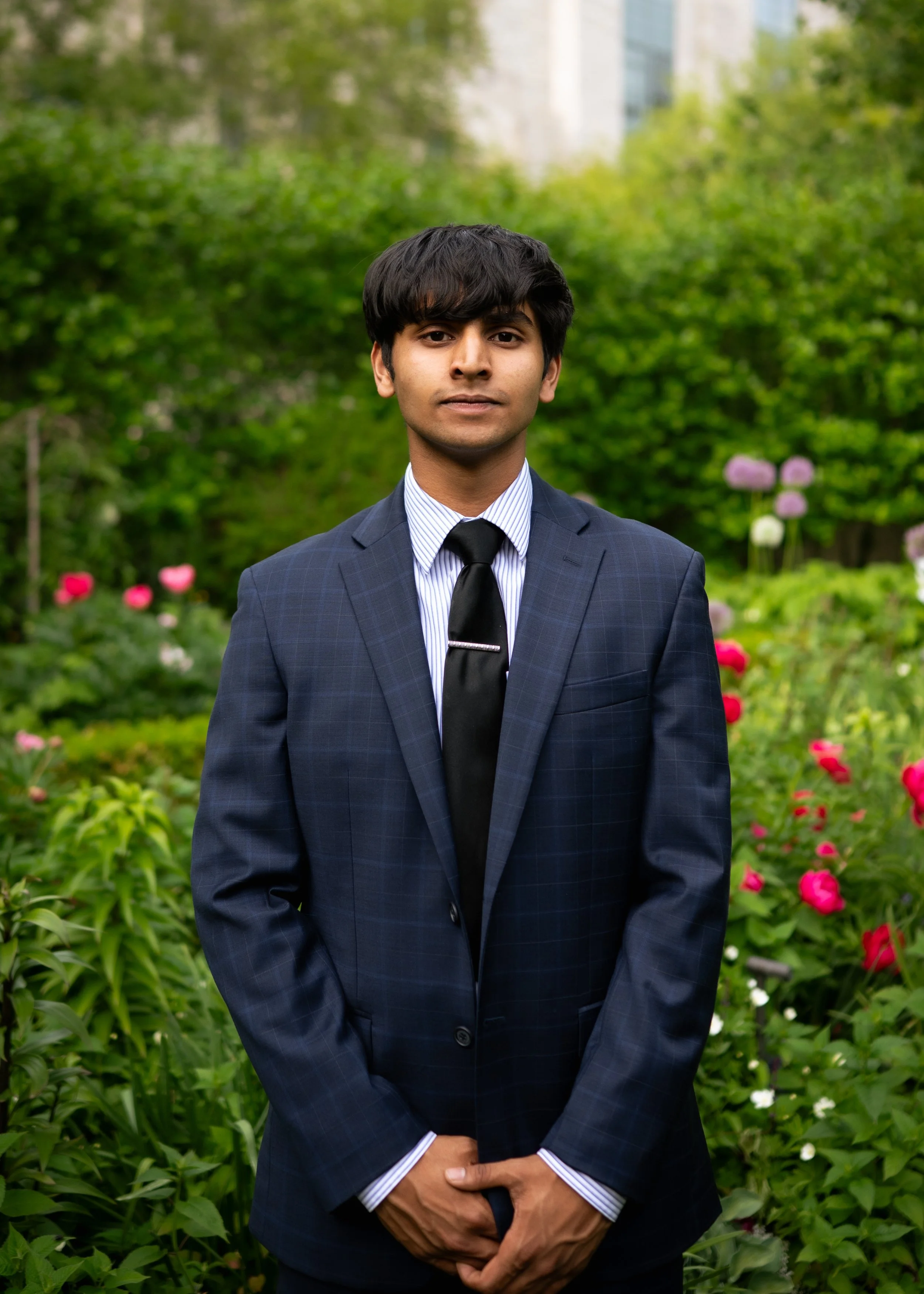 A young man wearing a dark business suit and tie stands outdoors with green bushes and pink flowers in the background.