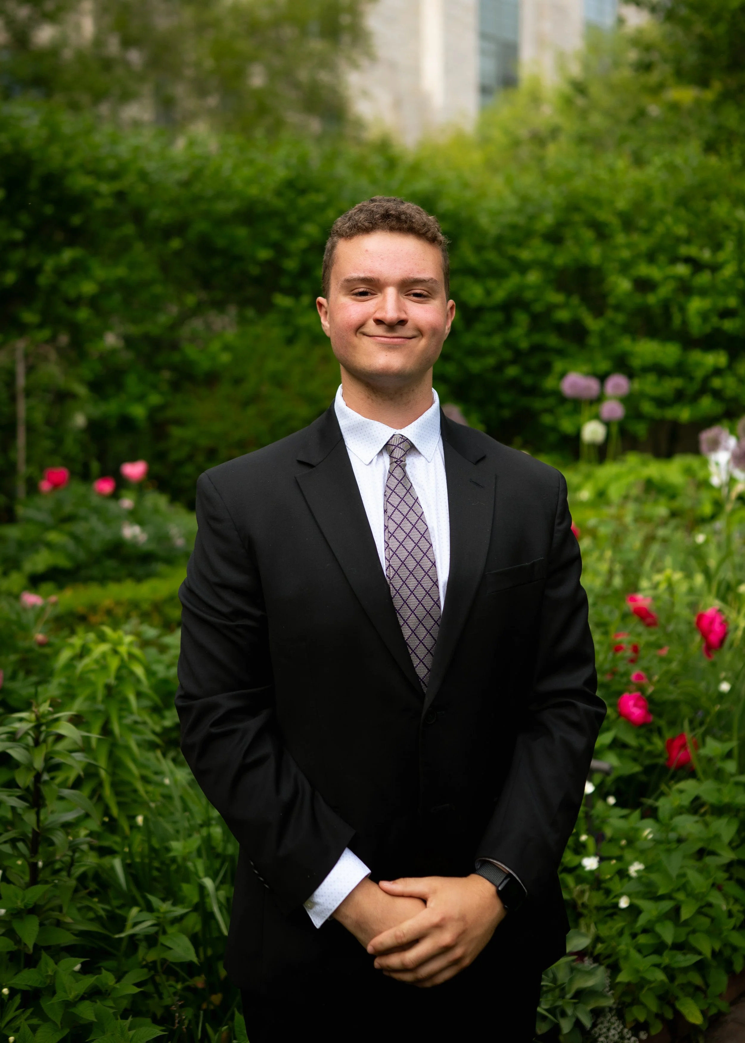 A young man dressed in a suit with a patterned tie stands outdoors in front of lush green foliage and colorful flowers, smiling at the camera.