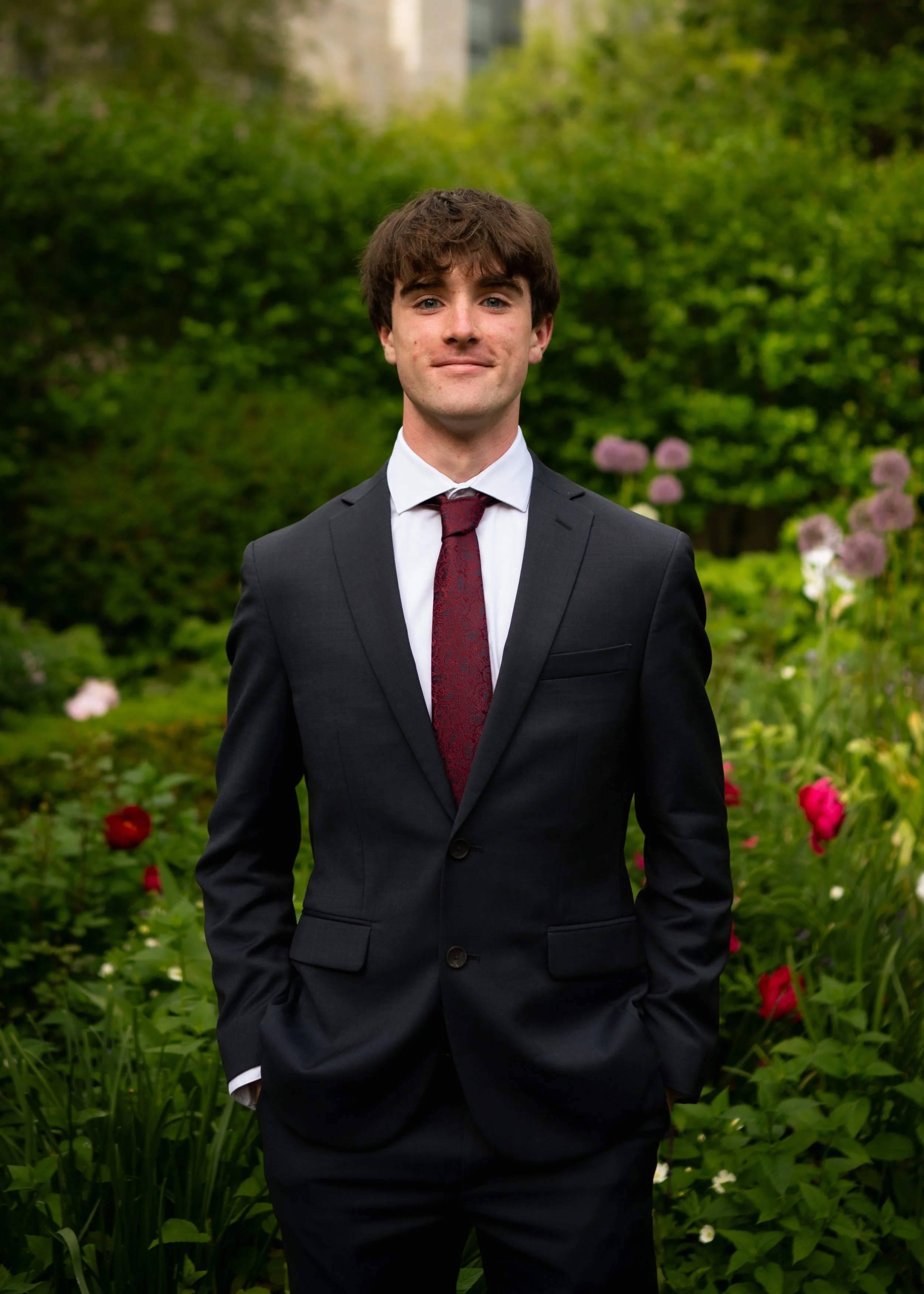 A young man in a dark suit with a white shirt and red tie standing outdoors in a garden with green foliage and pink flowers.