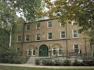 A historic four-story brick building with a green door and multiple windows, surrounded by trees and a sidewalk.