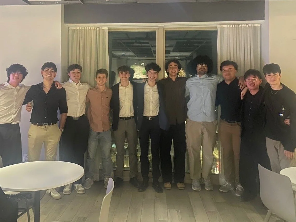 Group of young men standing together indoors at a social gathering, smiling, with some having their arms around each other, behind a window with closed curtains.