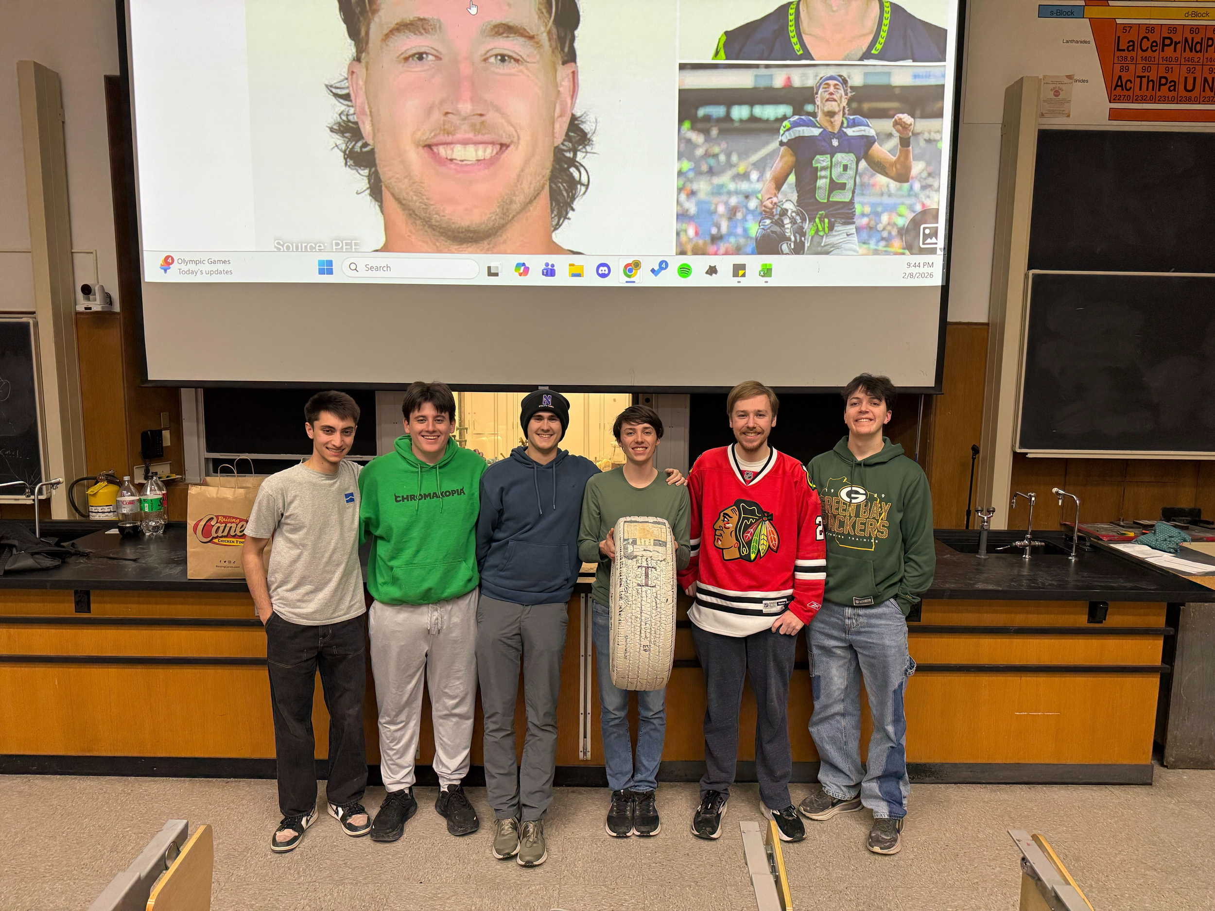 Six young men standing side by side in a classroom, smiling at the camera. They are dressed in casual sportswear and are holding a science or engineering project, which appears to be a tire or wheel with some written notes on it. Behind them, there is a blackboard, some classroom supplies, and a large screen displaying images of a man and a football player.