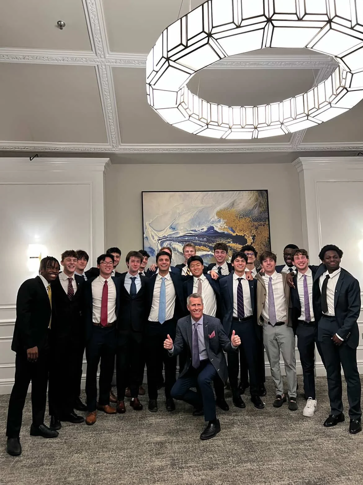 Group of young men in suits and ties, posing with an older man in a formal setting, all smiling and giving thumbs up.