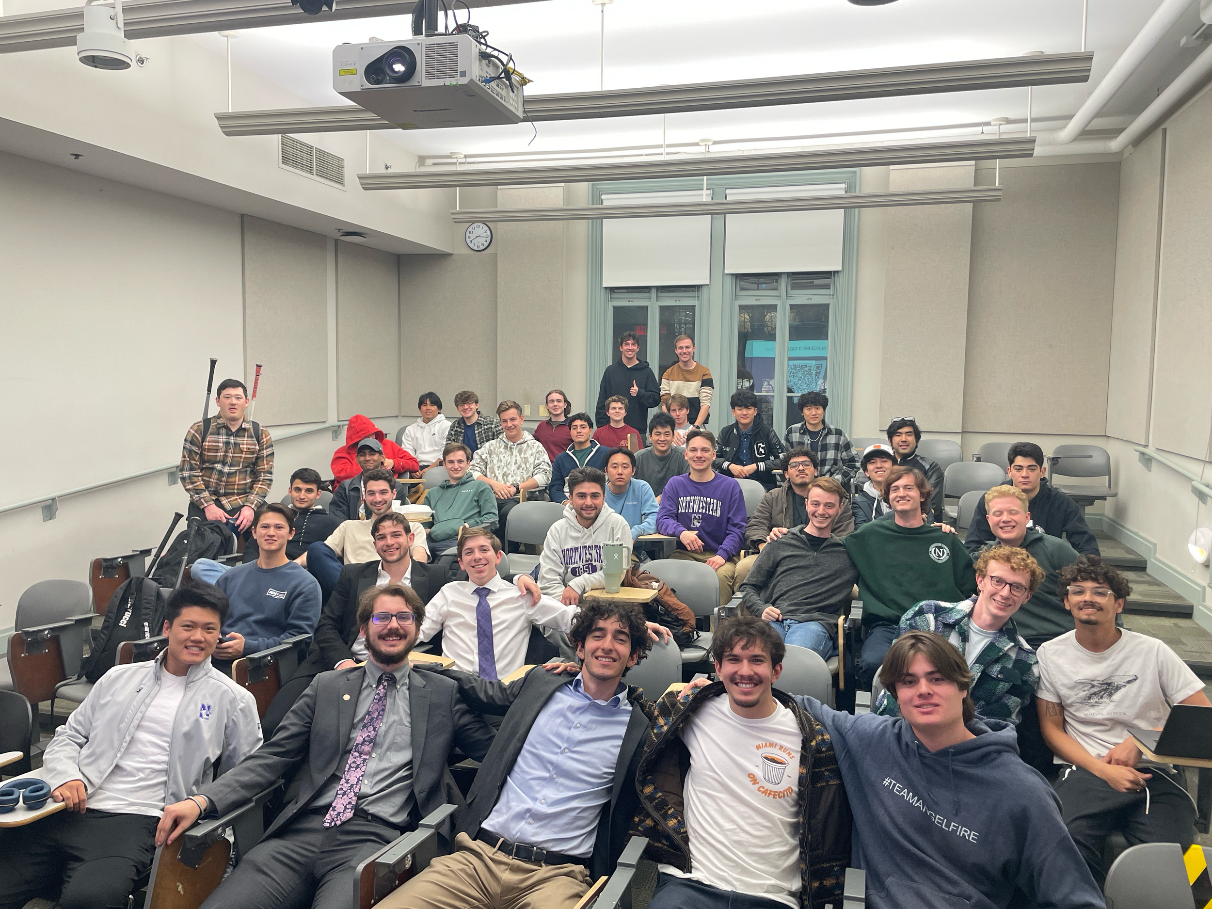 Group of students sitting in a lecture hall, smiling and posing for the photo, with some students making gestures like thumbs up.