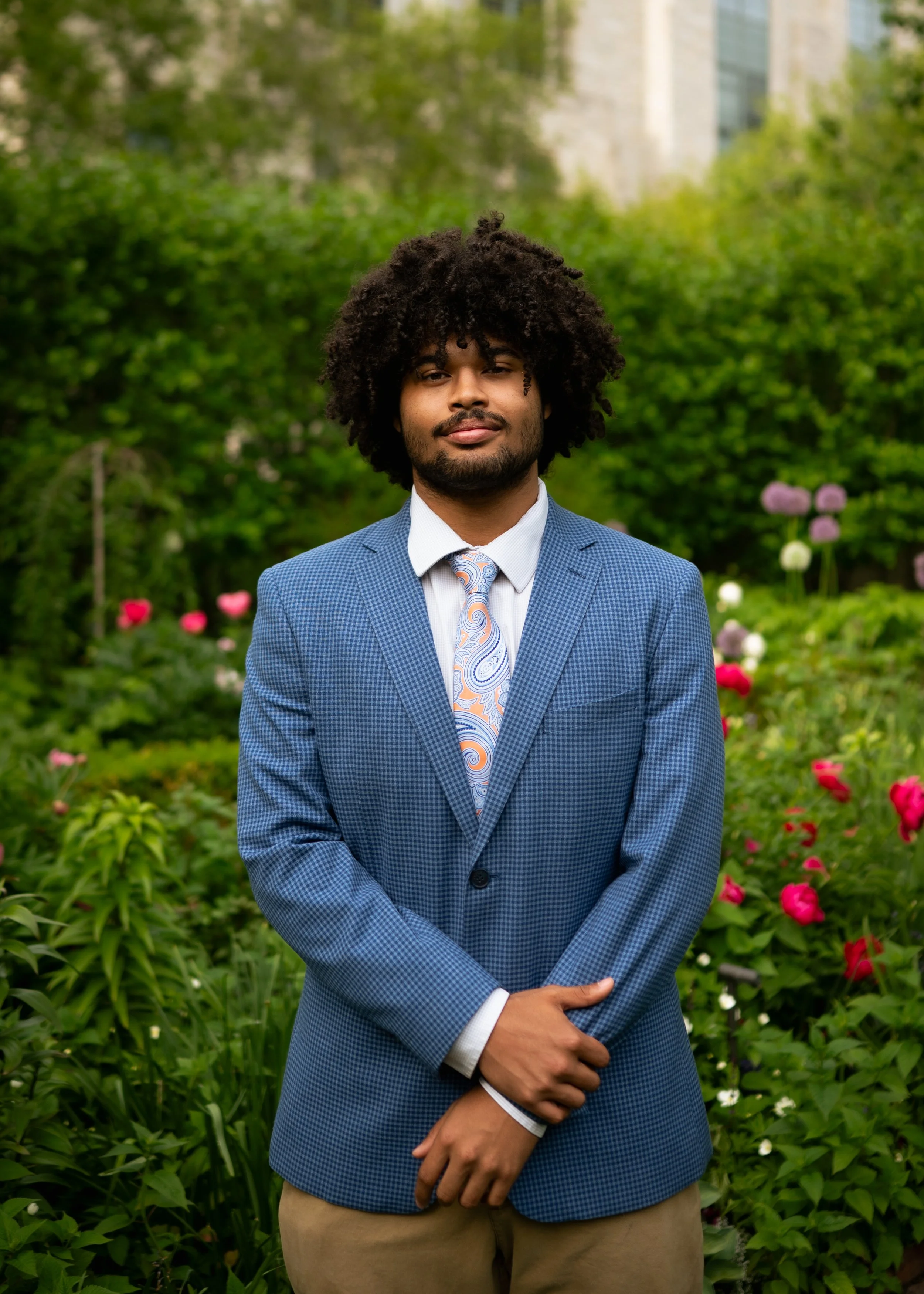 A man with curly hair and a beard, wearing a blue checkered blazer, white shirt, and patterned tie, standing outdoors in front of a lush green garden with flowers and trees.