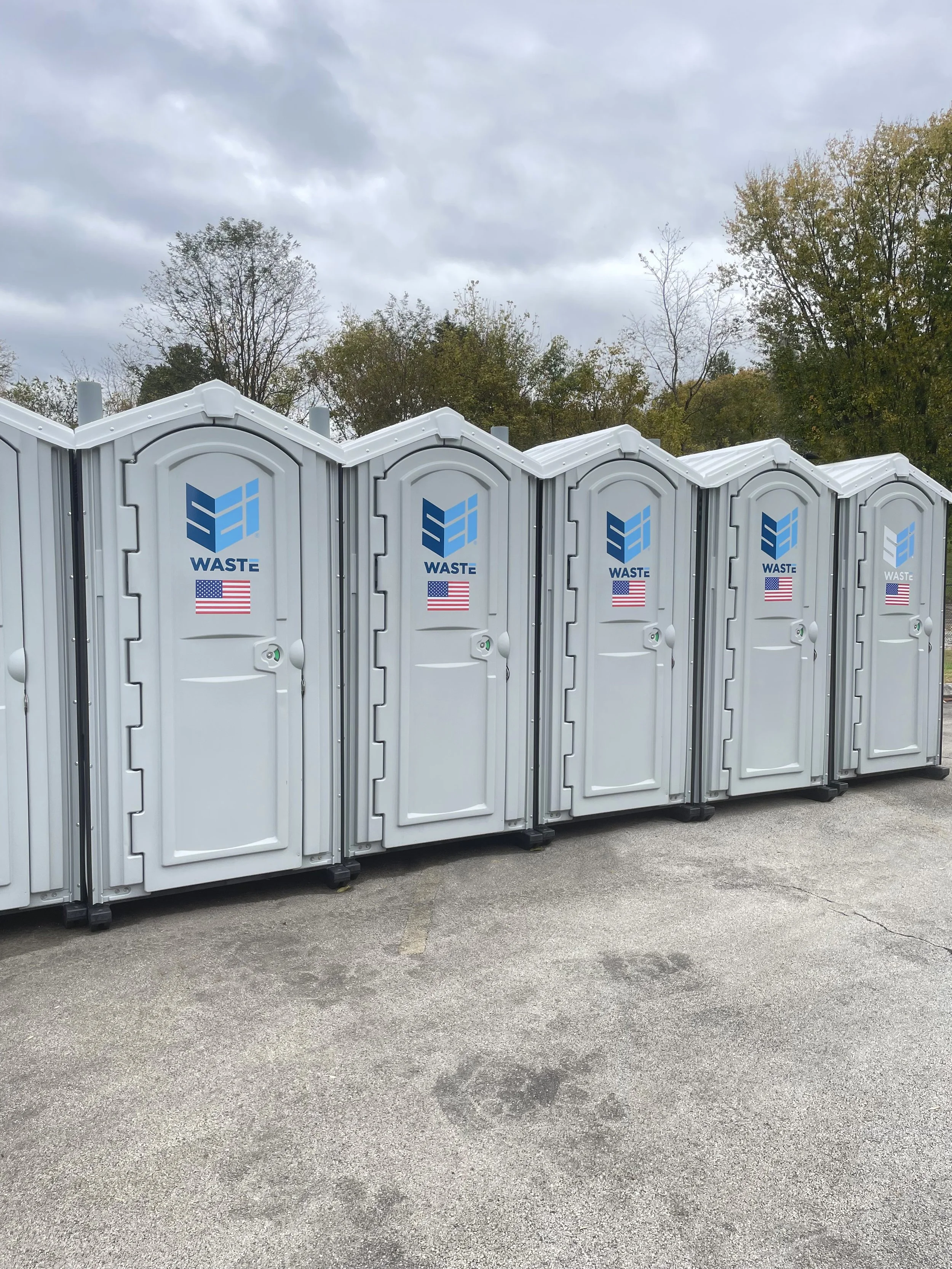 A row of SEI Waste rental portable toilets with blue waste recycling logos on the doors, set against a background of trees and overcast sky.
