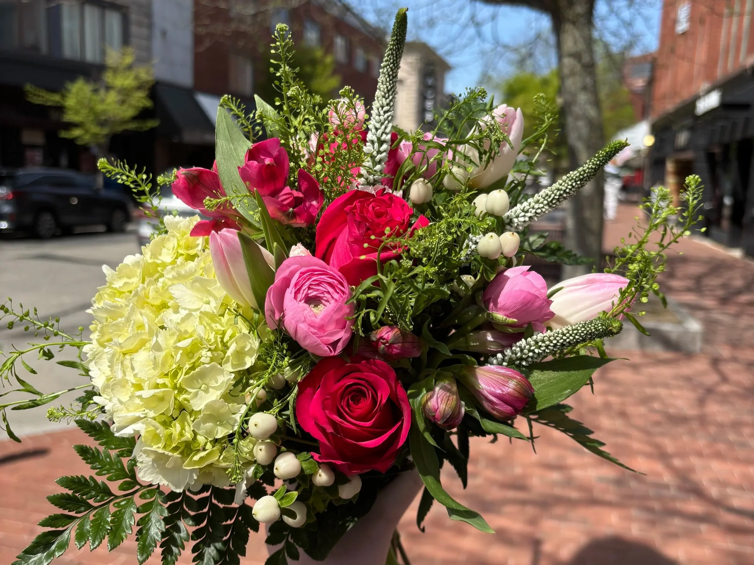 A vibrant bouquet of mixed flowers including pink roses, tulips, white berries, and greenery being held outdoors on a sunny day with a city street background.