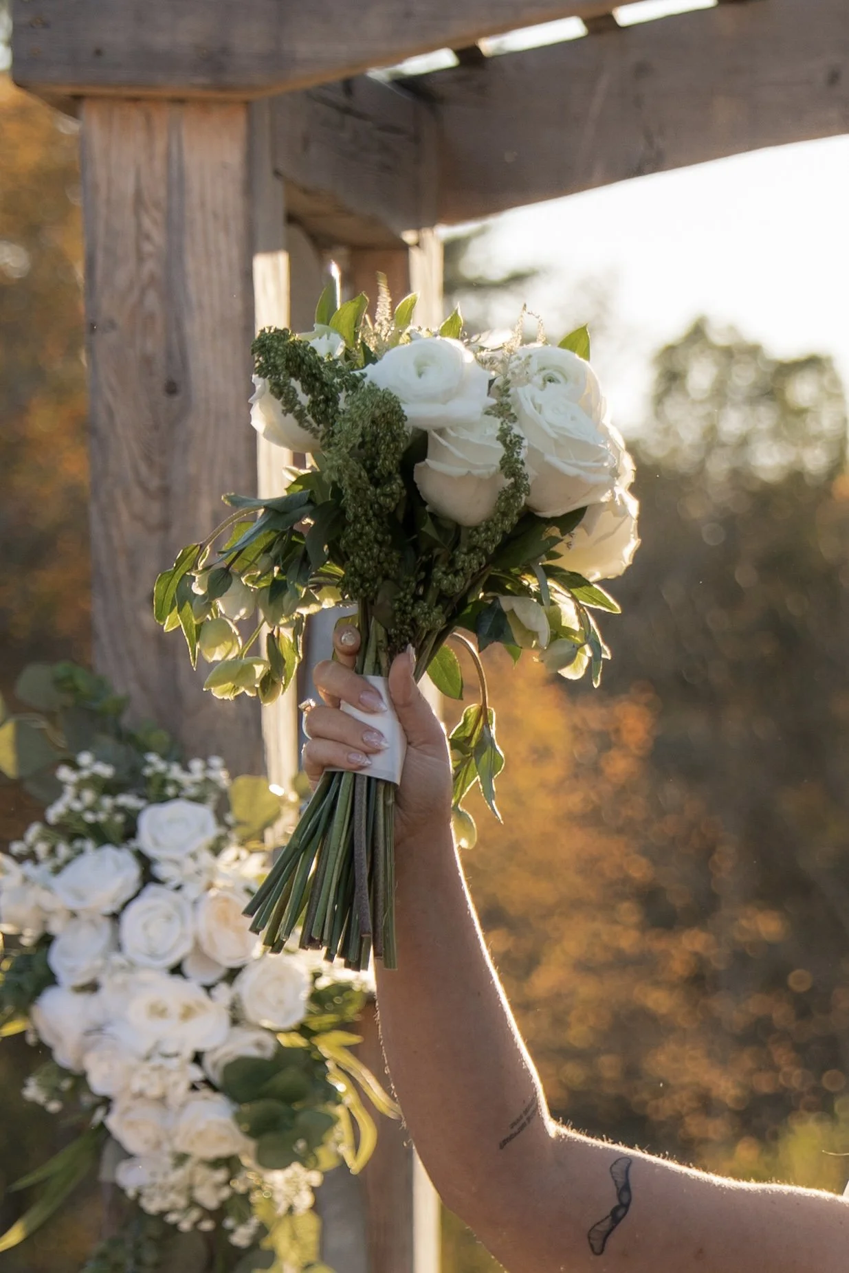 A hand holding a bouquet of white roses and greenery outdoors during sunset, with a wooden structure and blurred trees in the background.