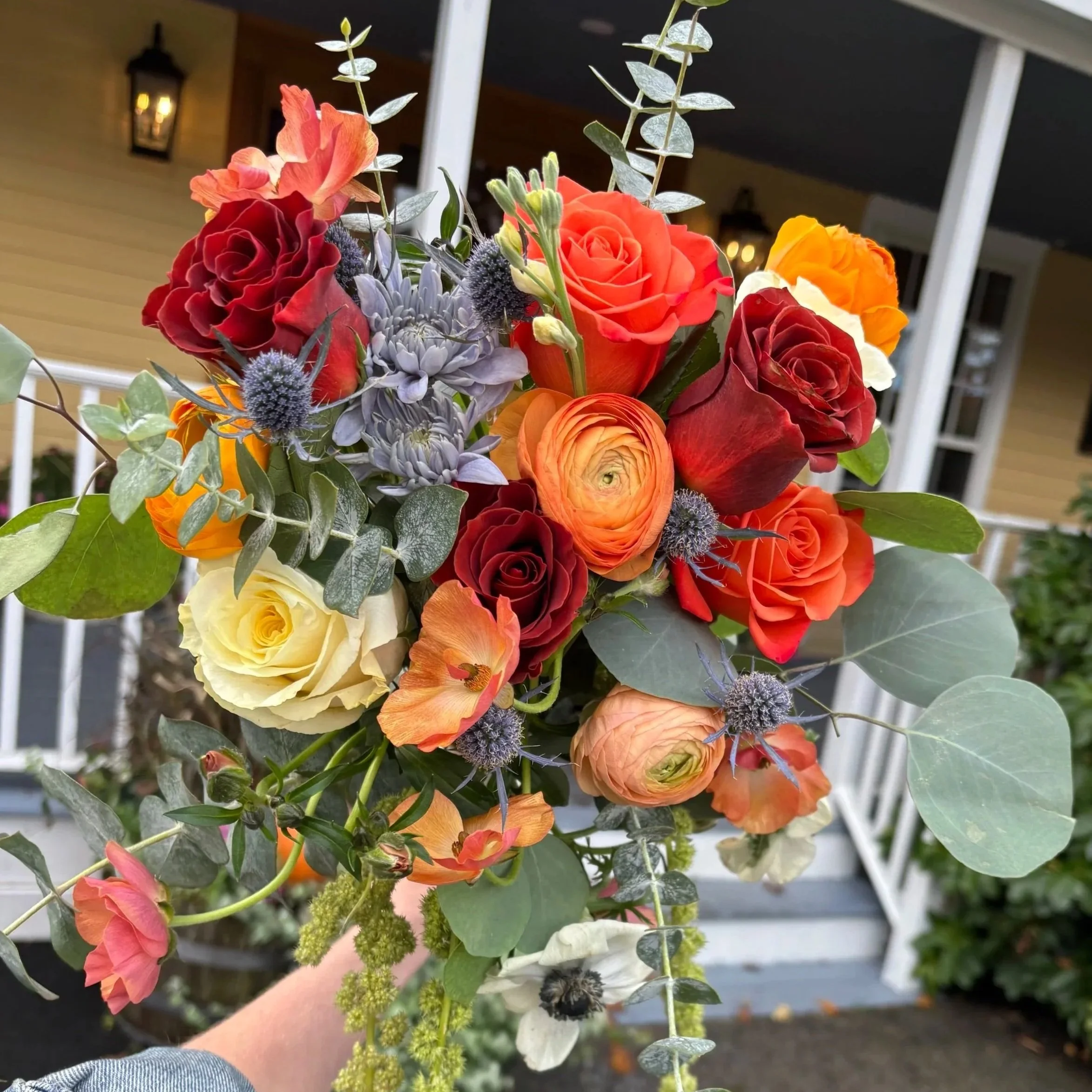A colorful bouquet of flowers including roses, ranunculus, and various greenery, held up in front of a house porch with a walkway and a lantern light.