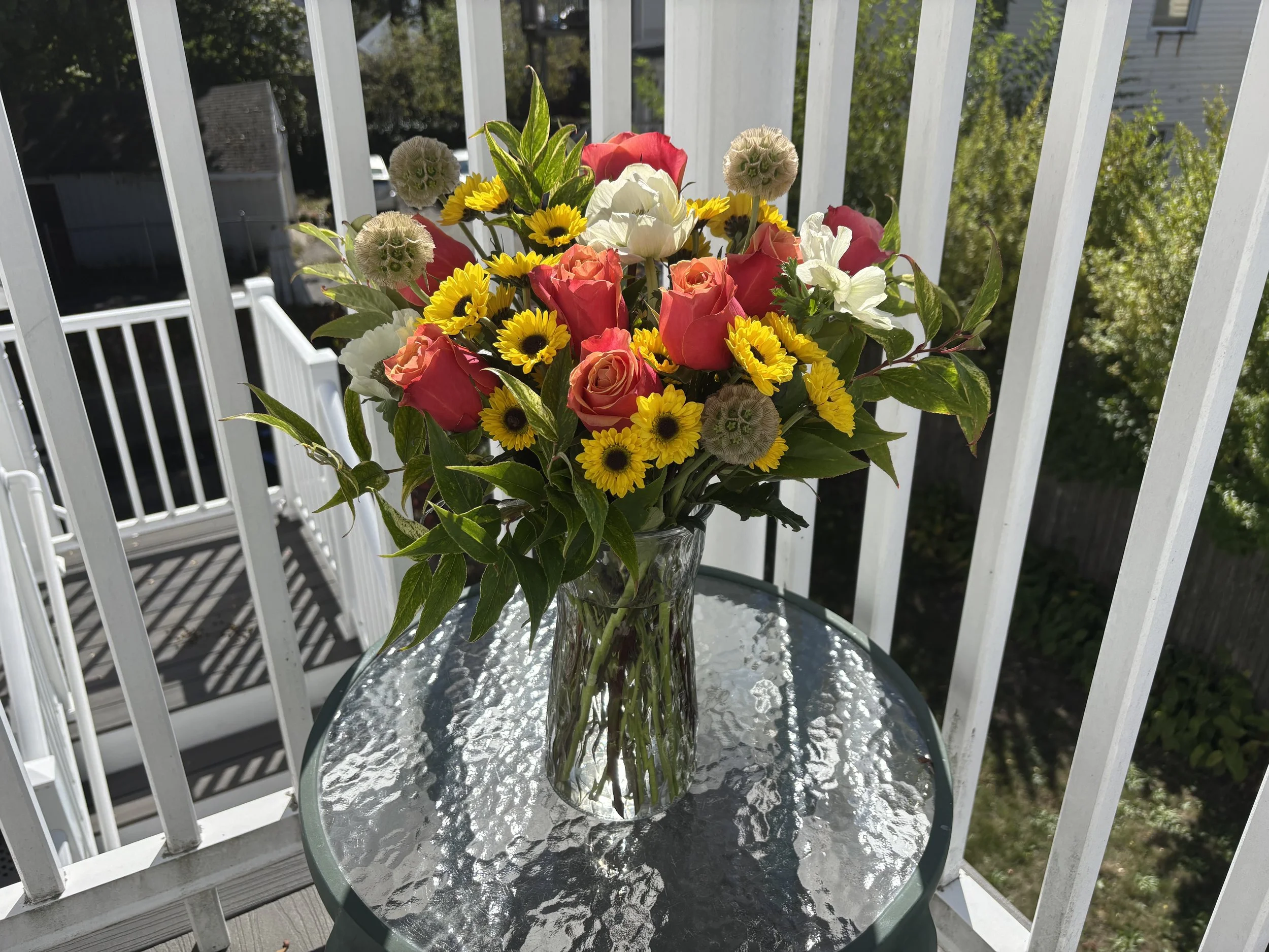 A glass vase filled with a colorful bouquet of pink roses, white flowers, yellow daisies, and green leaves, placed on a small glass-topped table on a balcony with white railing.