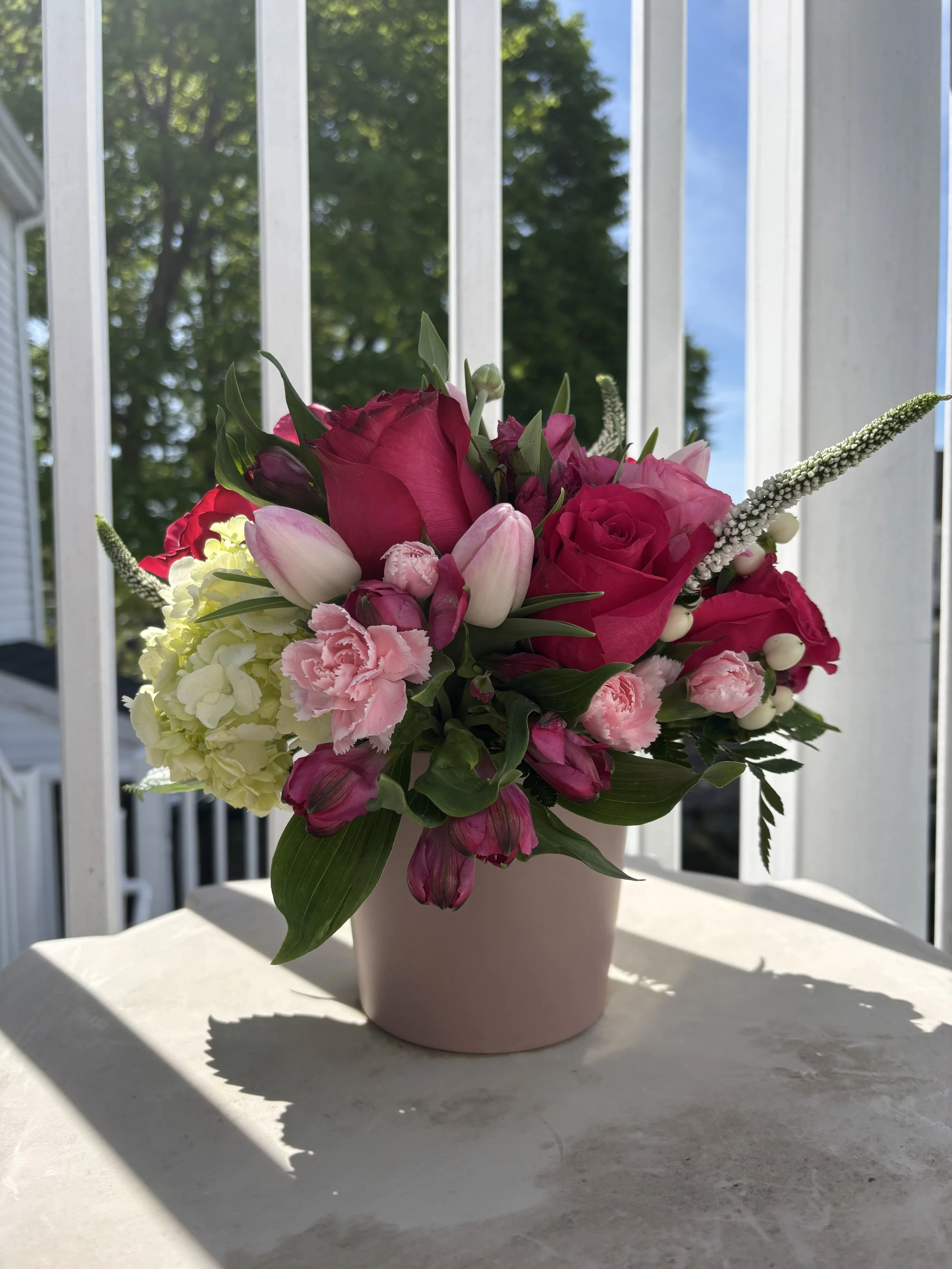 A pink flower arrangement in a pink vase on a table, outdoors, with trees and blue sky in the background.