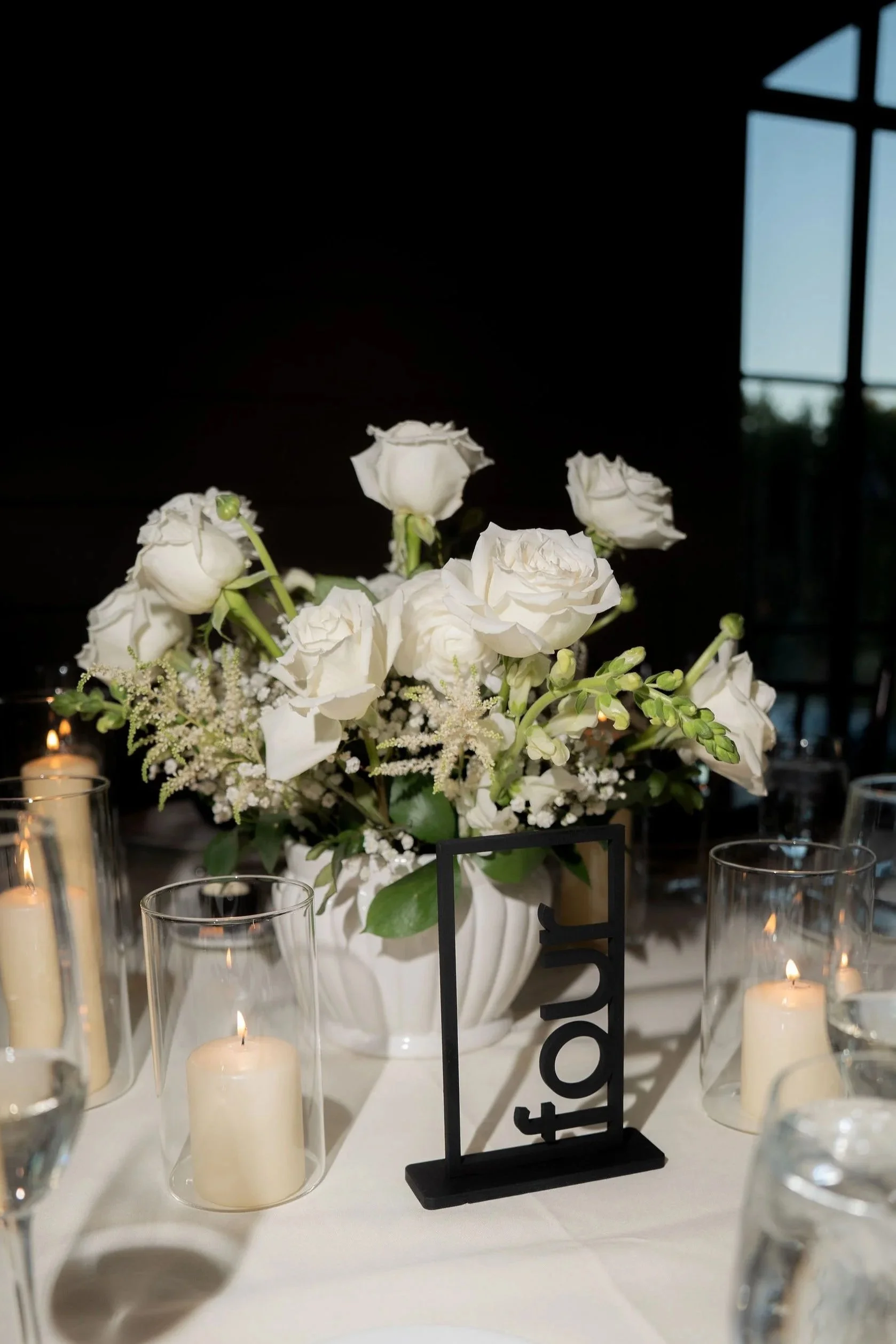 White floral centerpiece with candles on a table for a wedding or event, with a small black table number sign.