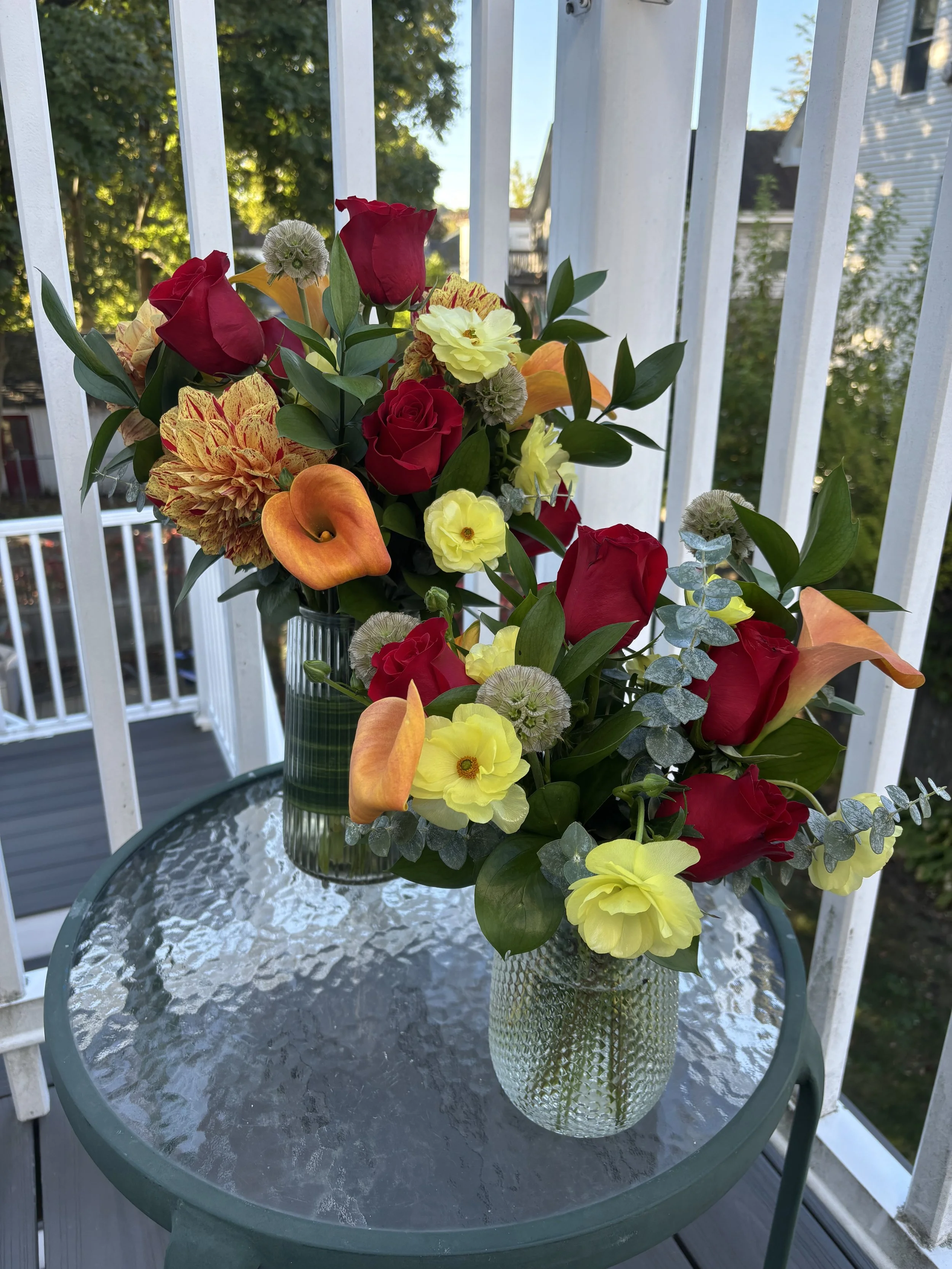 A glass table with two vases of colorful flower arrangements, featuring red roses, orange calla lilies, yellow primroses, and dried seed heads, on a balcony with white railings, trees, and houses in the background.