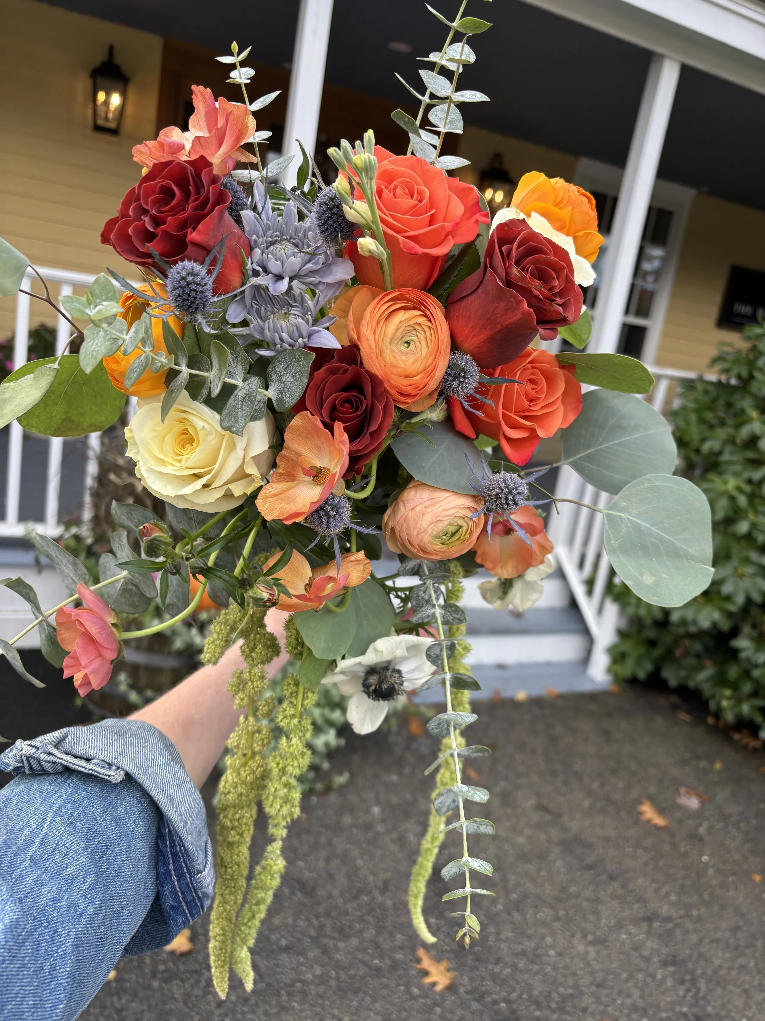 A hand holding a vibrant flower bouquet outside a house with a porch, featuring orange, red, yellow roses, purple thistles, and green foliage.