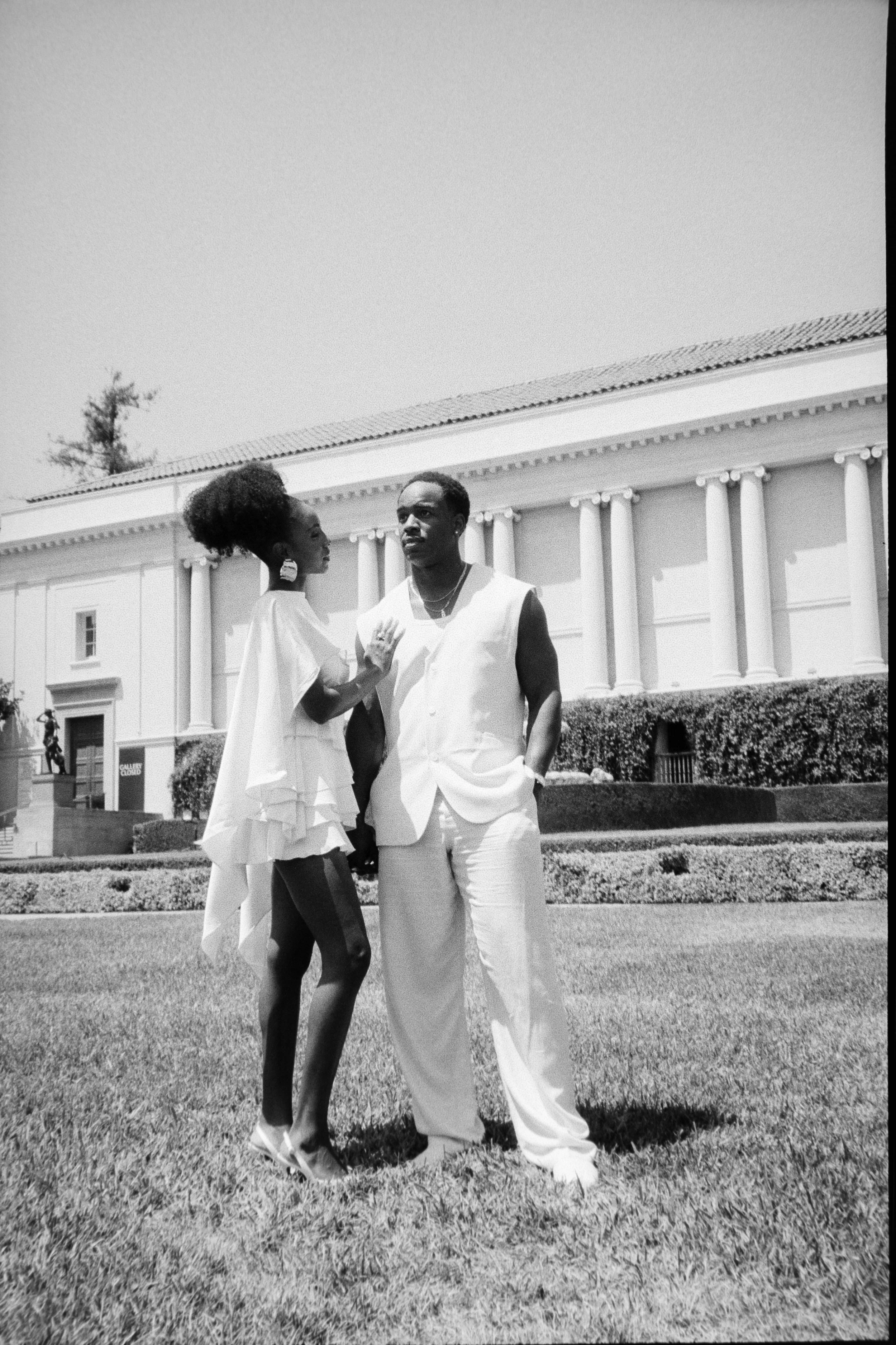 A black and white photo of a woman and a man standing on a grassy area in front of a large, classical-style building with columns. The woman has her hand on the man's chest, and both are looking in opposite directions. The woman has curly hair and is